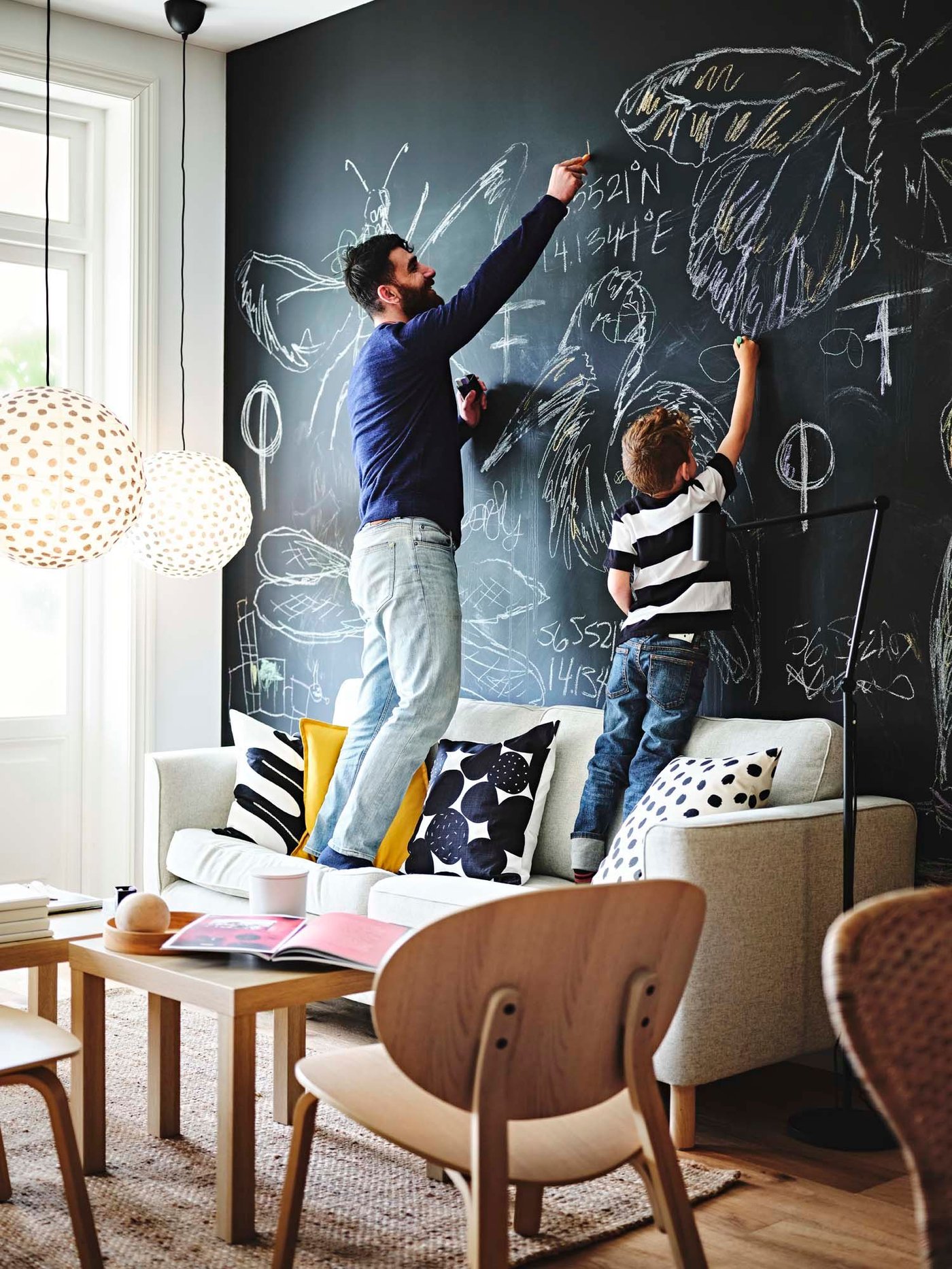 A man and his son stand on a sofa, they are drawing with chalk on a blackboard wall behind the sofa.