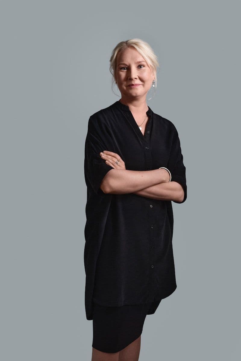 Designer Iina Vuorivirta stands smiling with folded arms against a light grey backdrop.