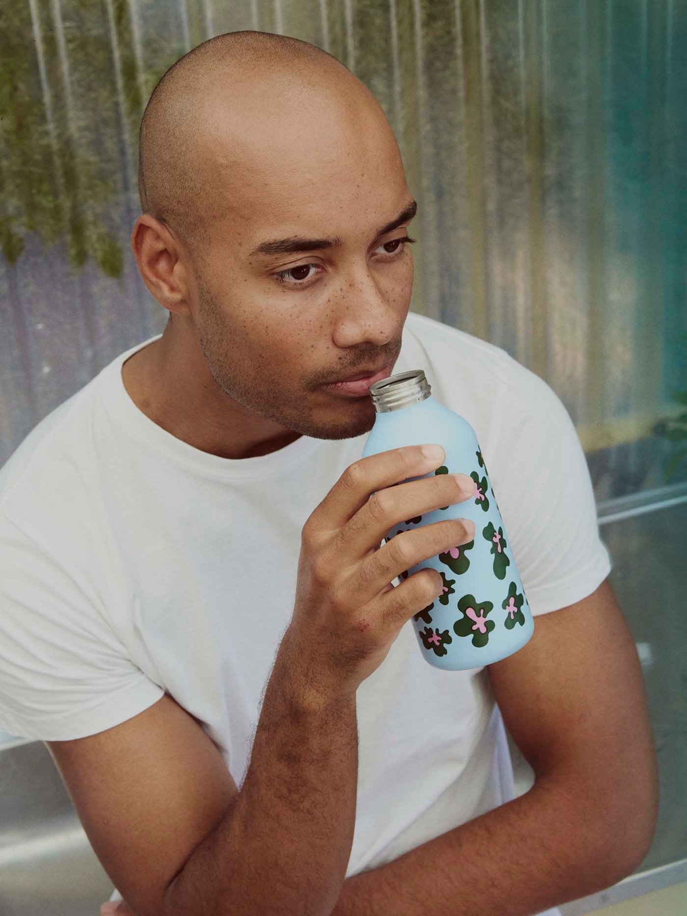 A man sits. He is drinking from a patterned water bottle from the BASTUA collection.