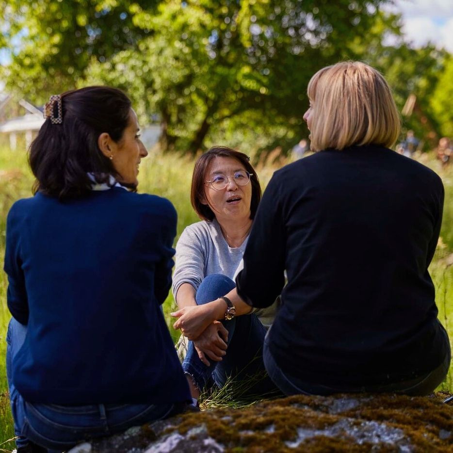 Three people sit together on the grass. Two of them have their back to the camera.