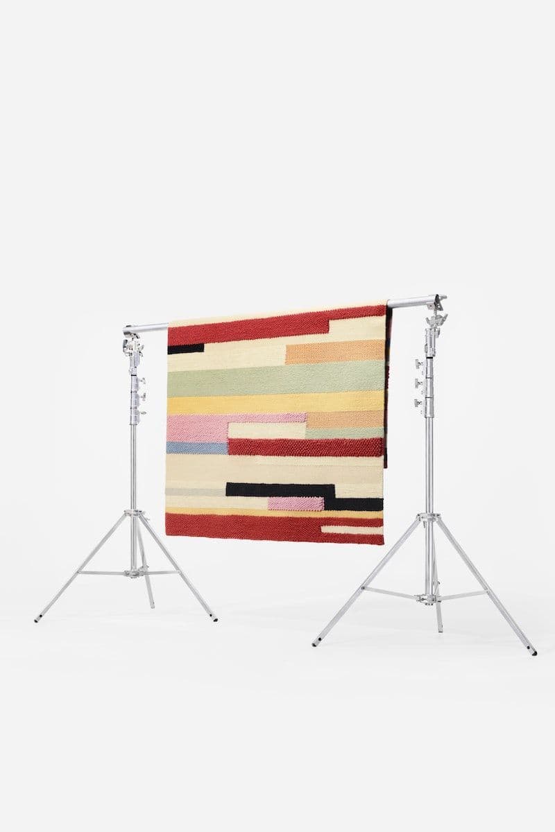 A colourful BRÖNDEN rug hangs from a metal frame in a white studio setting.