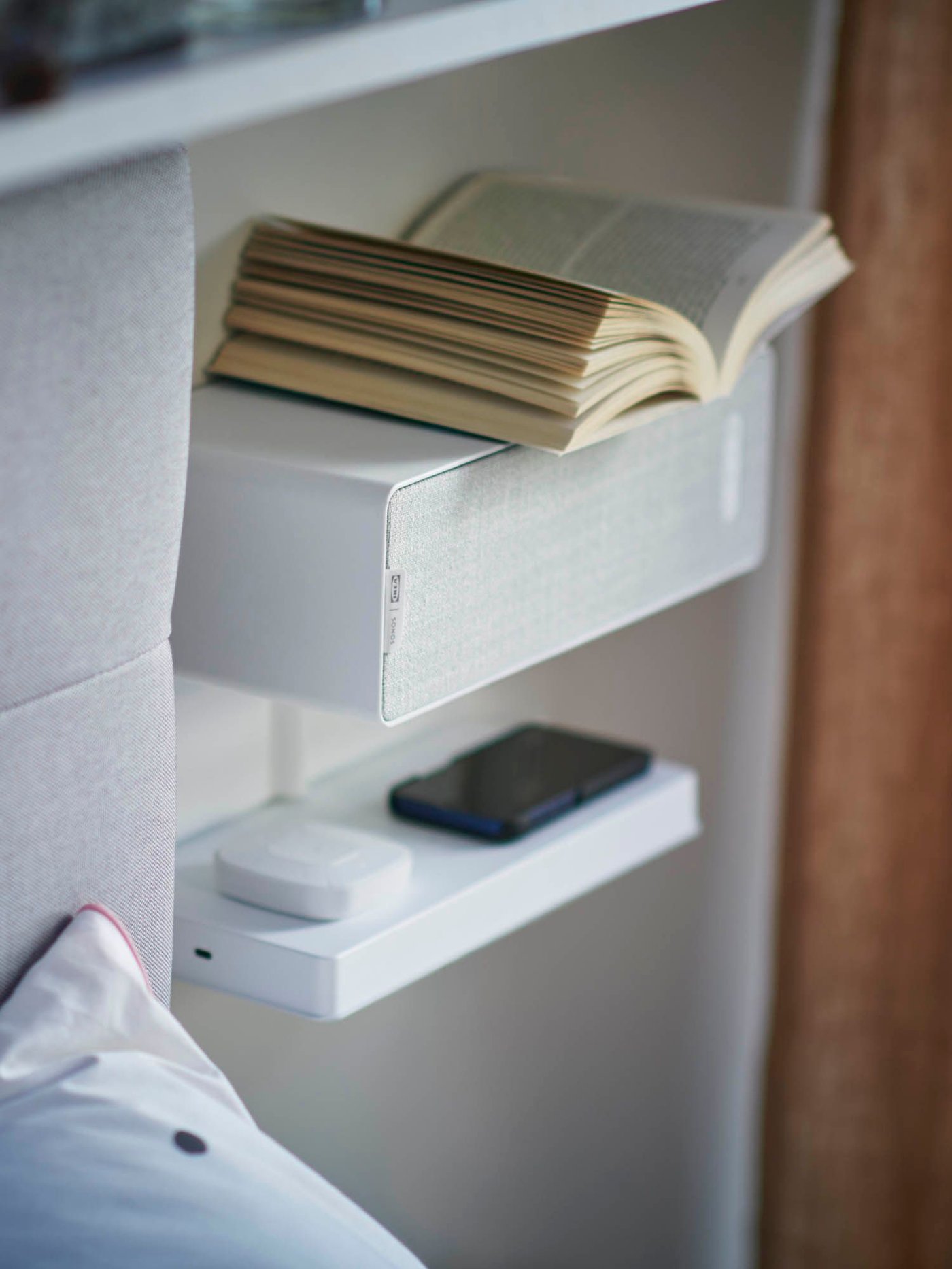 Next to the edge of a headboard, a shelf and a SYMFONISK Wifi bookshelf speaker are attached to the wall. An open book sits on top of the bookshelf speaker.
