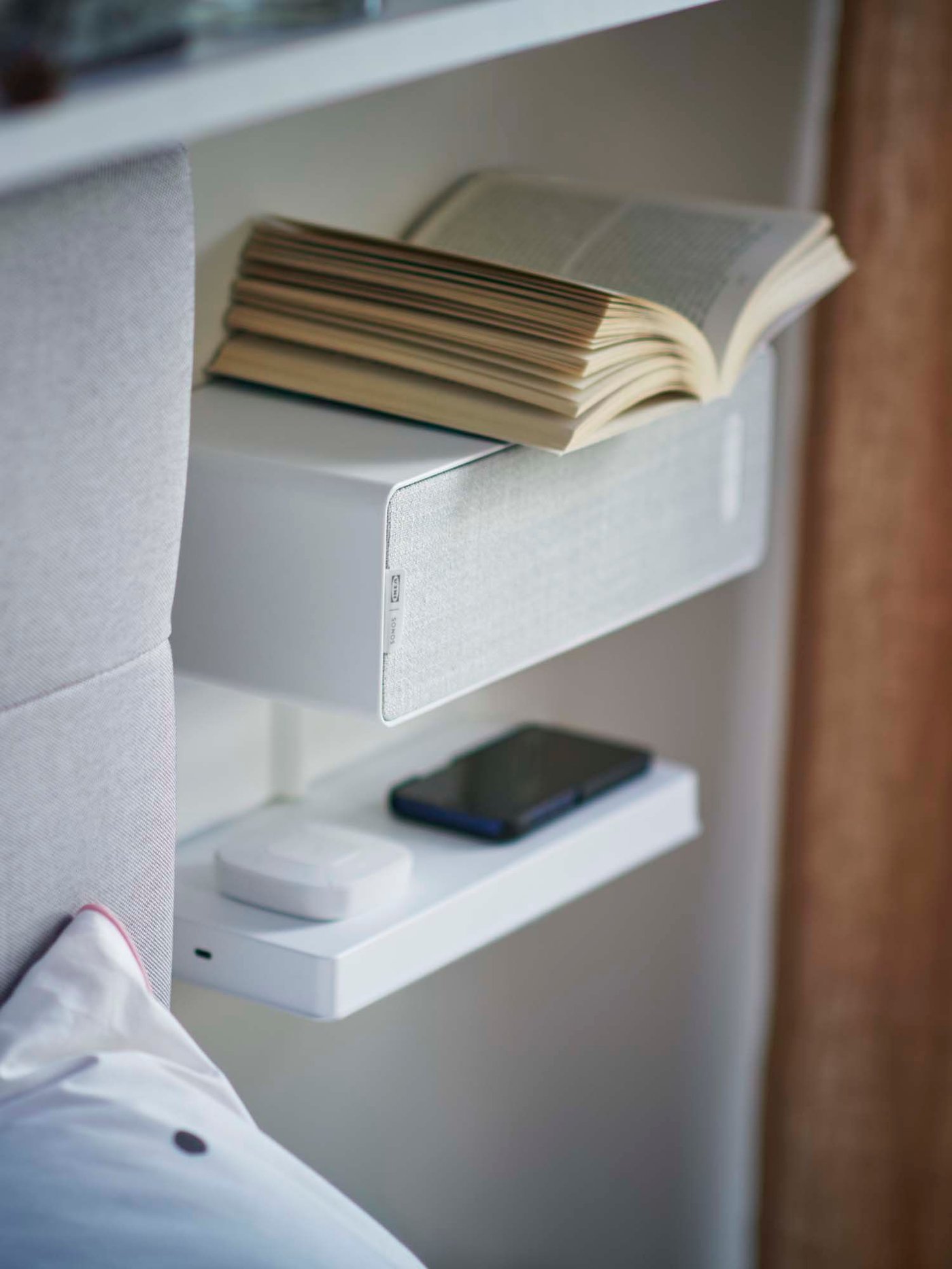 Next to the edge of a headboard, a shelf and a SYMFONISK Wifi bookshelf speaker are attached to the wall. An open book sits on top of the bookshelf speaker.