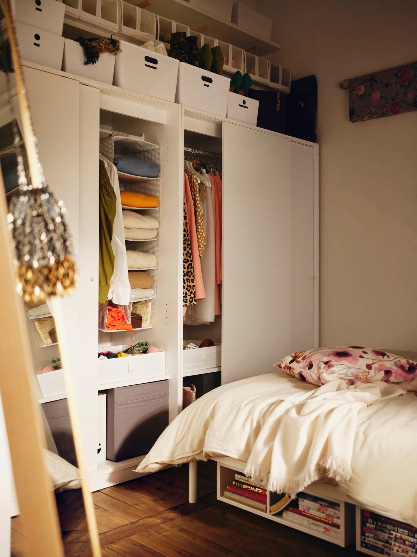 The corner of a bedroom with a large wardrobe. The doors are open revealing shelves and hanging clothes. There are boxes above.