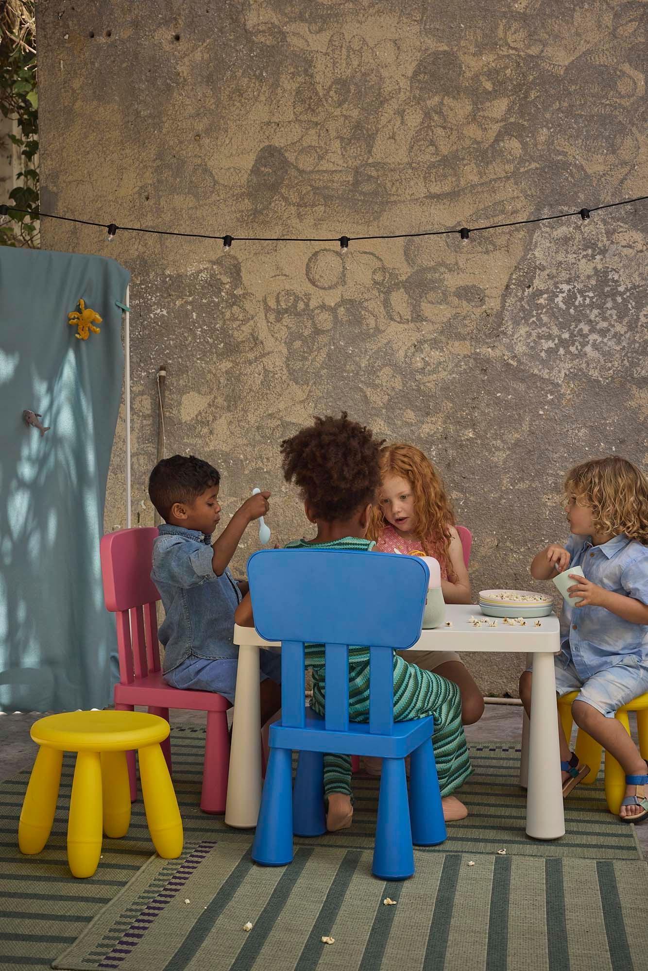 Children sitting at a white MAMMUT table with MAMMUT chairs and stools in pink, yellow and blue, on a green rug.