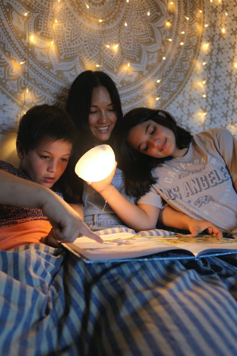 A woman and two children sit in bed, reading a bedtime story. The girl holds a SAMMANLÄNKAD light in her hand, shining light on the book. Stars are projected on the wall behind them.