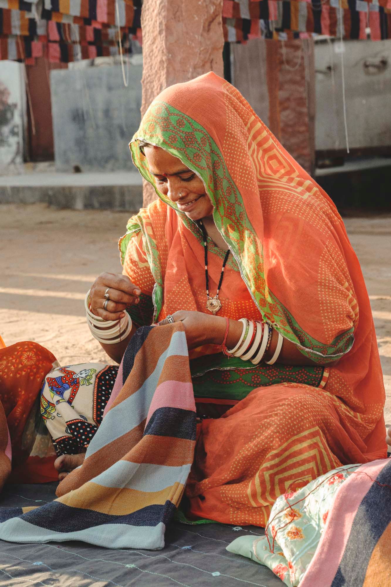 A woman sits on the floor embroidering. She is smiling.