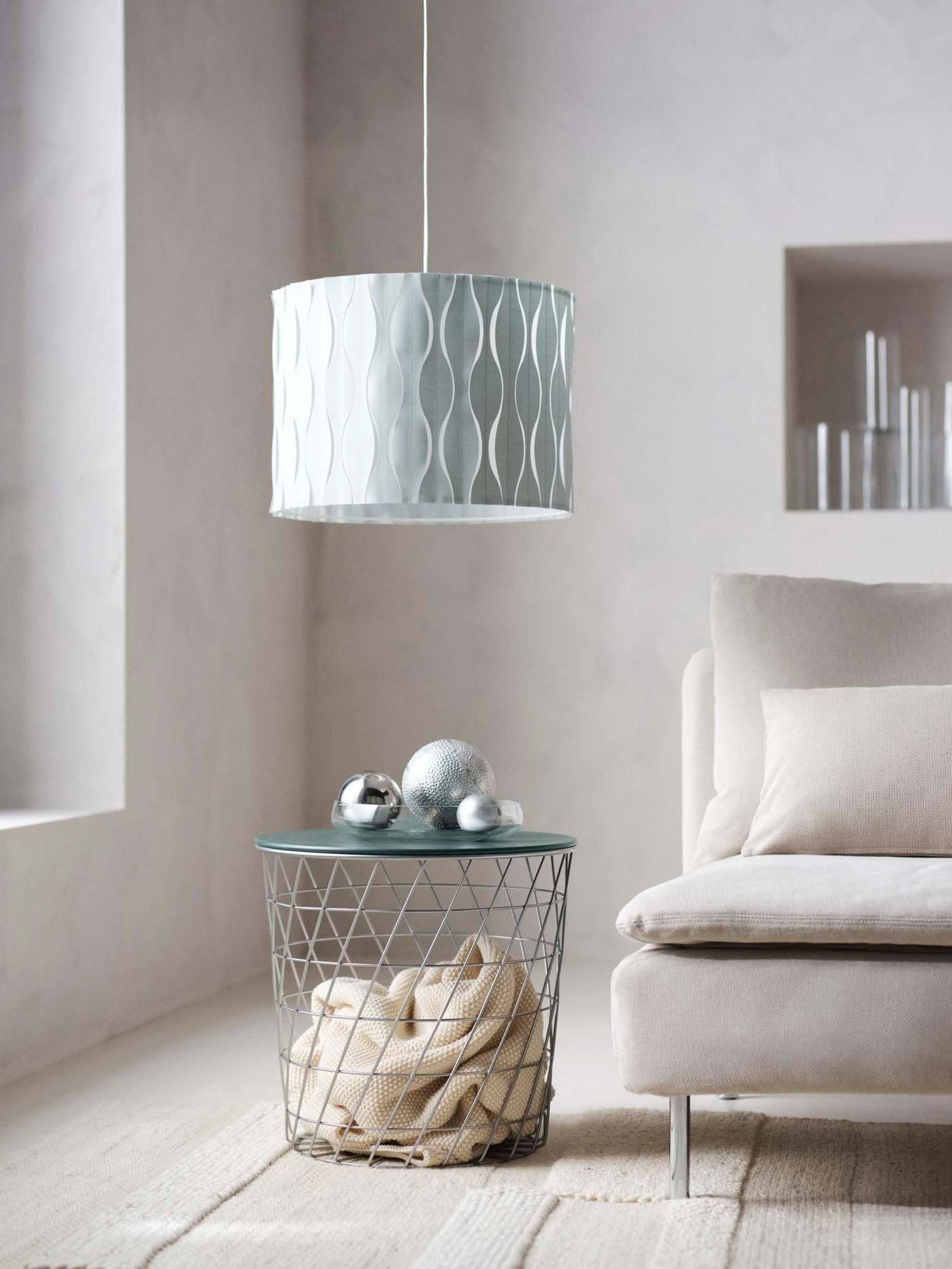 The corner of a pale living room. A light blue lampshade hangs over a KVISTBRO storage table, next to a grey sofa.