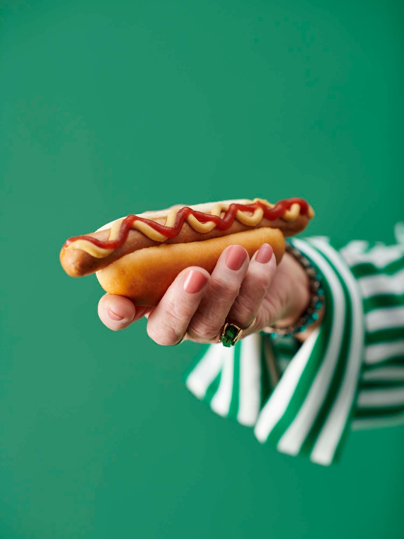 The hand of a person in a green striped shirt holds a hot dog.