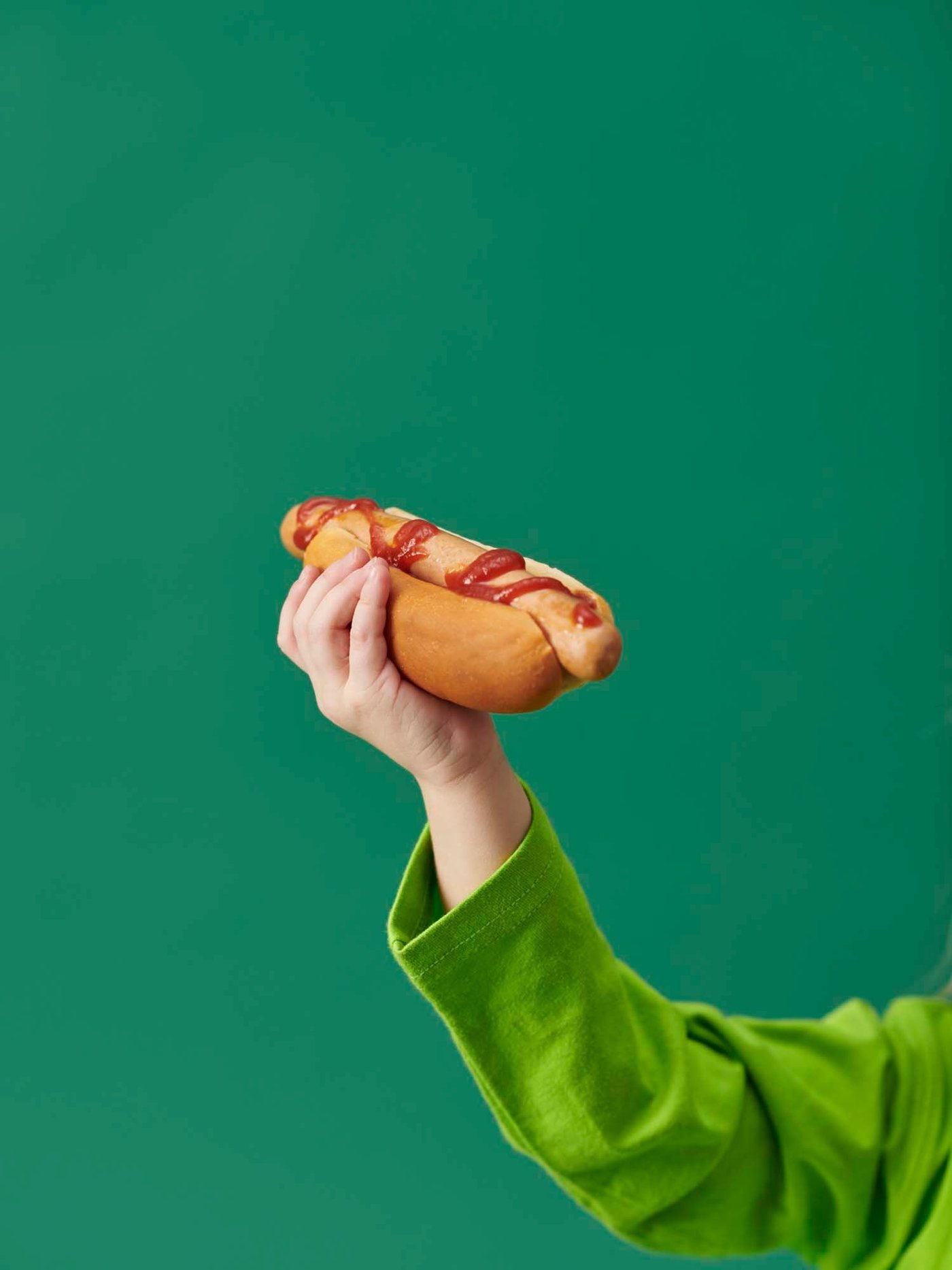 A person wearing a green jumper holds a hot dog in a bun high. The hot dog has ketchup and mustard on it.