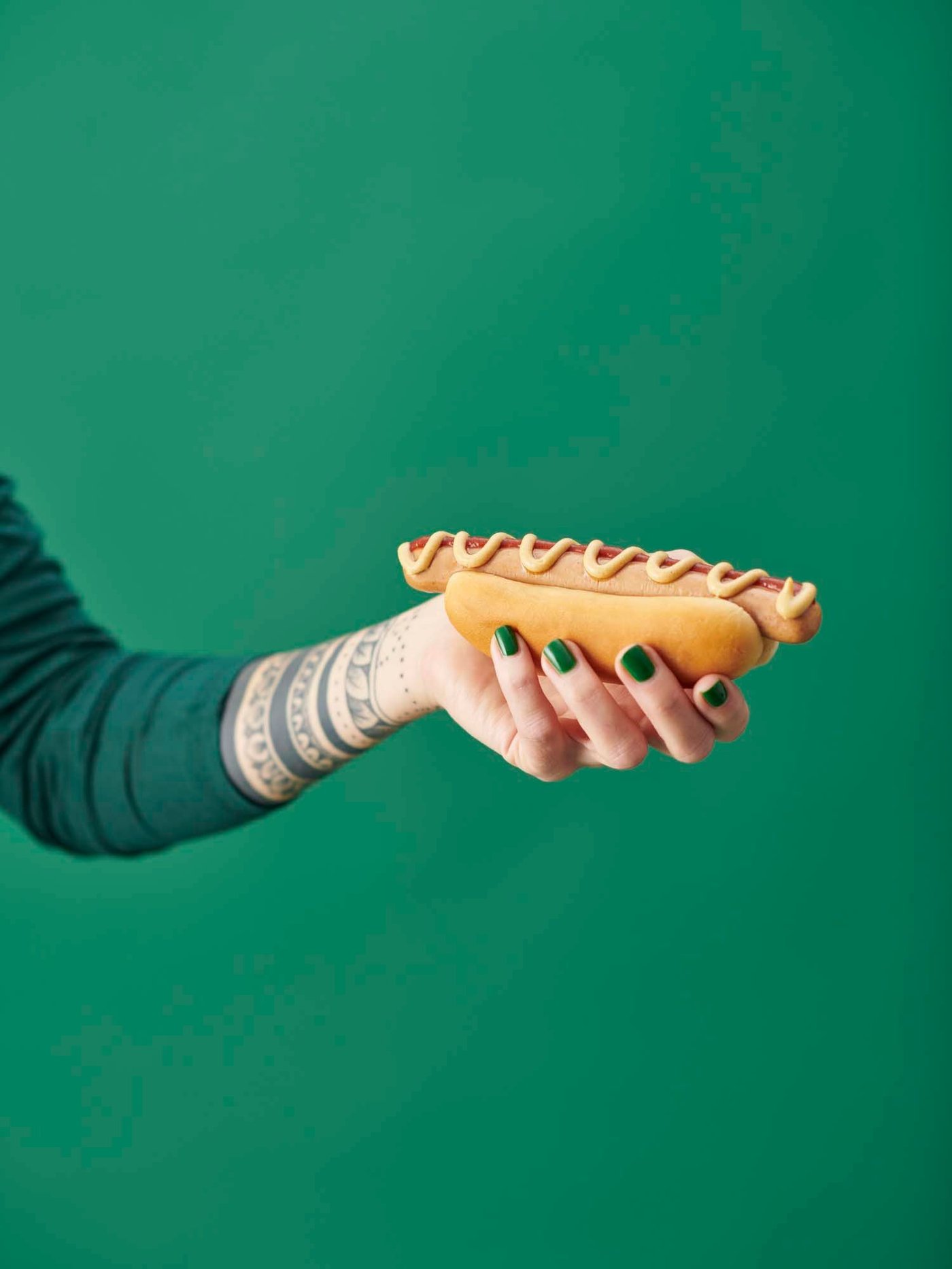 A hand with green nail polish holds a plant-based hot dog with ketchup and mustard.