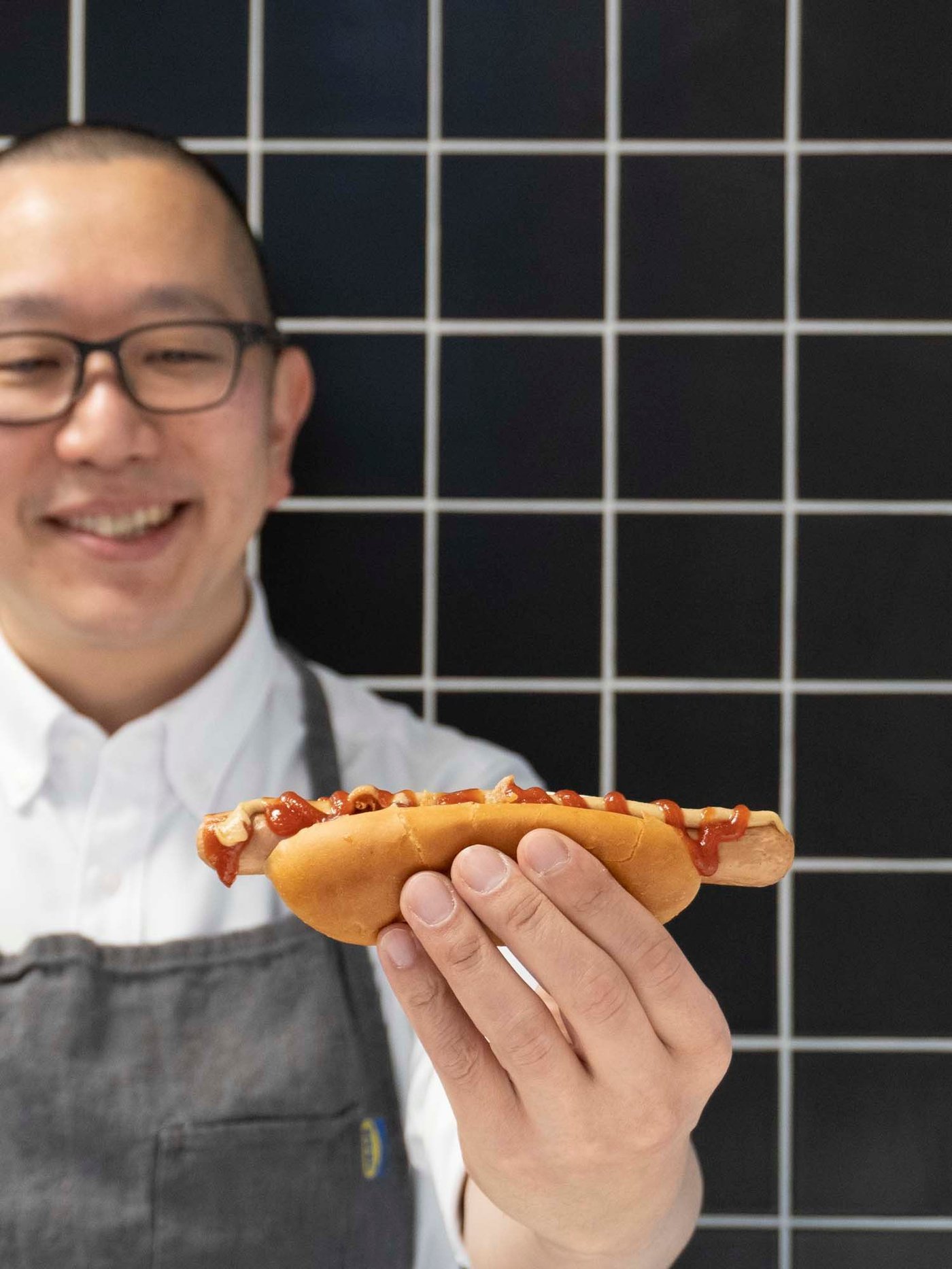 A man in an apron holds a plant-based hot dog up. There is a black tiled wall behind him.