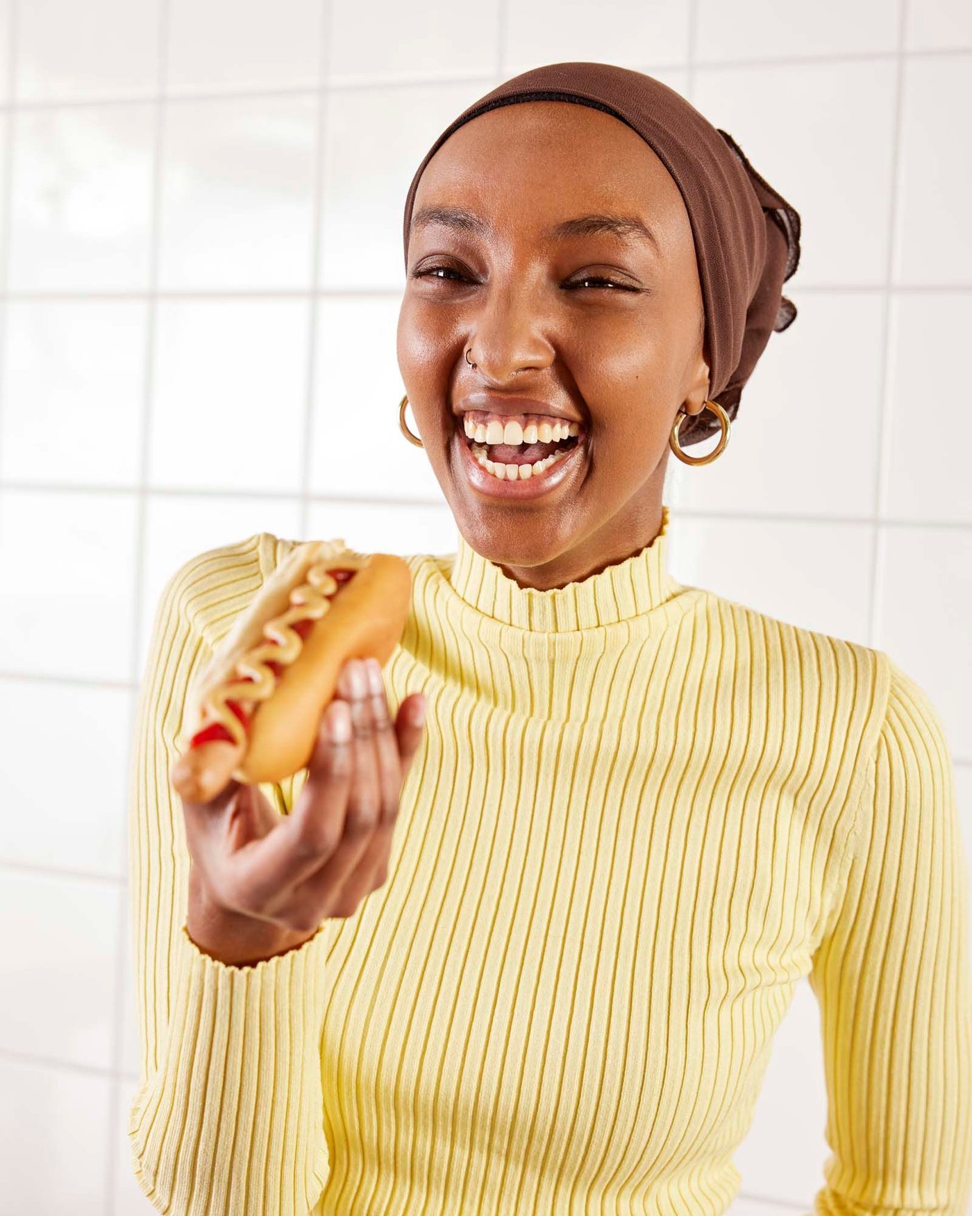 A young woman is eating a plant-based hot dog with mustard and ketchup. She is laughing.