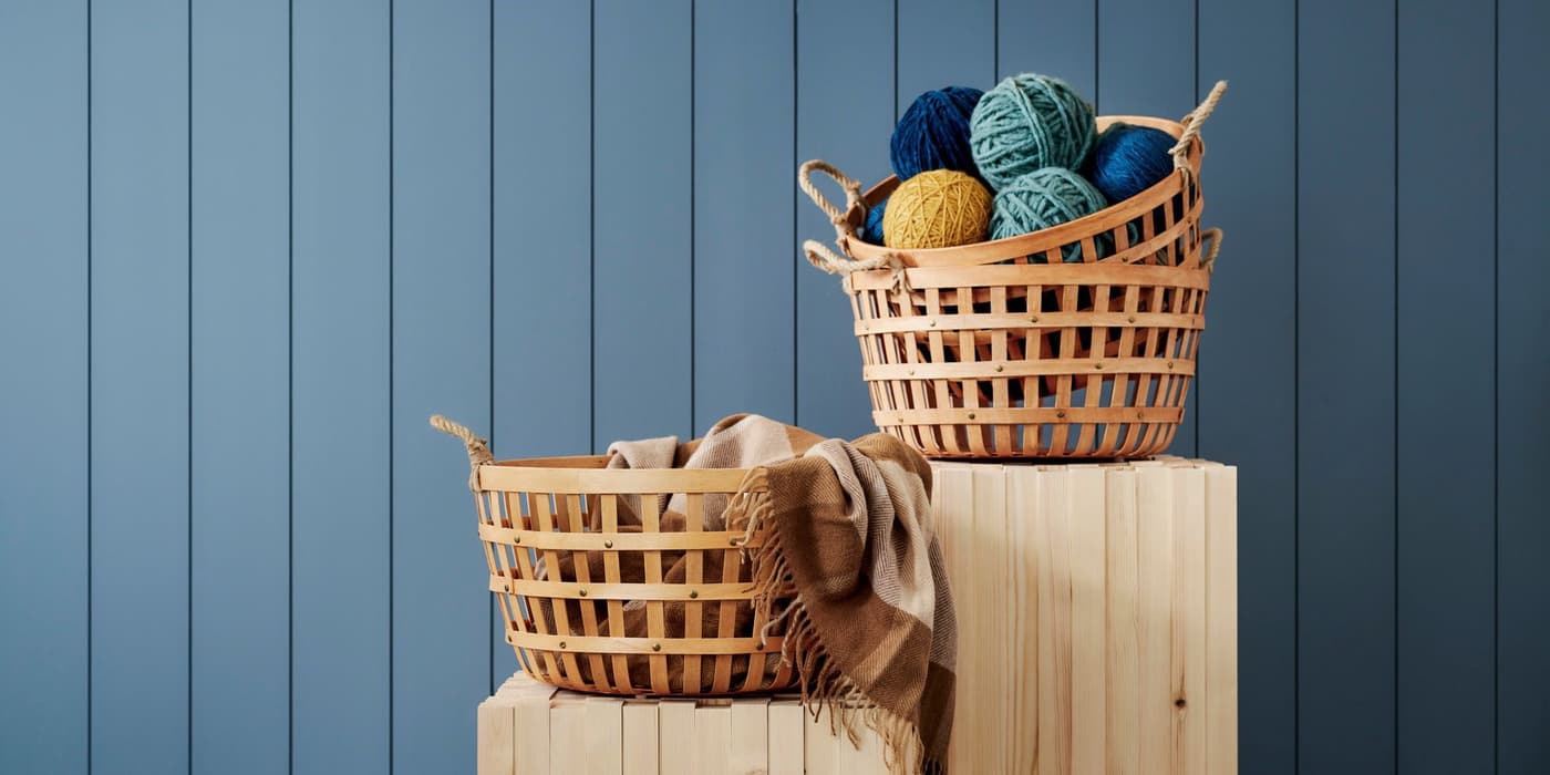 VÄXTHUS baskets filled with balls of colourful yarn, displayed on wooden plinths against a blue-painted, wood-panelled background. 