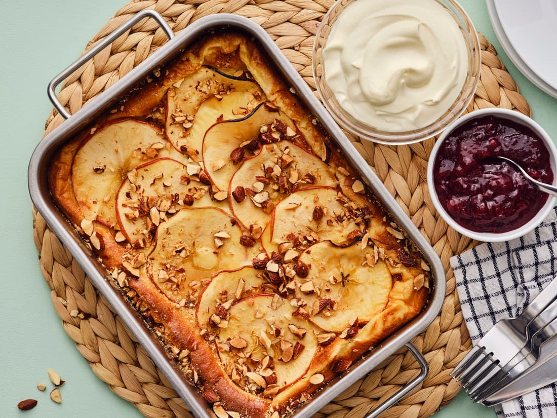 Apple topped pancake next to a bowl of jam and cream on a rattan trivet, on a pale green surface.