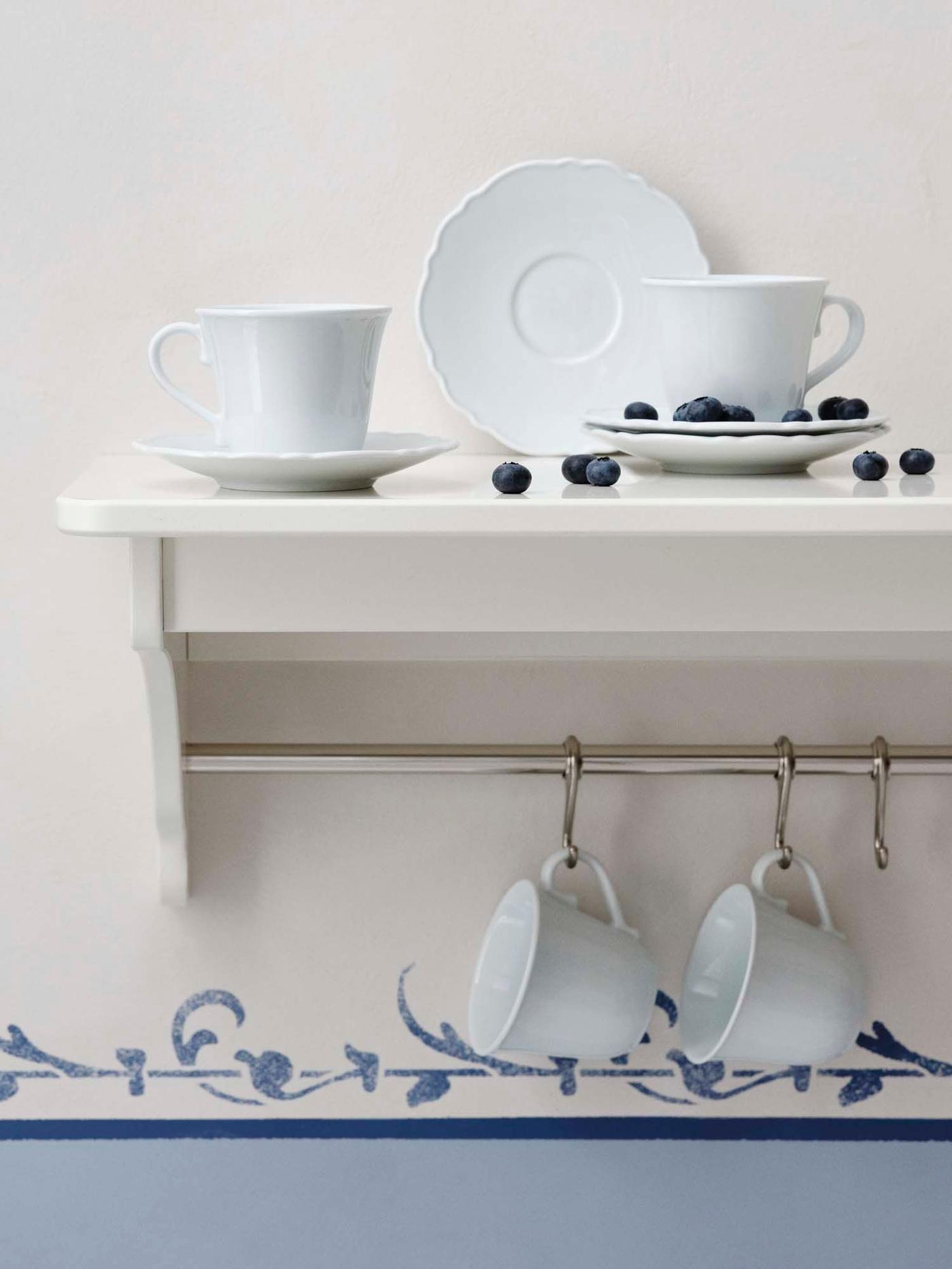 A white shelf attached to a blue and cream wall holds cups and saucers. Two more cups hang from hooks below.