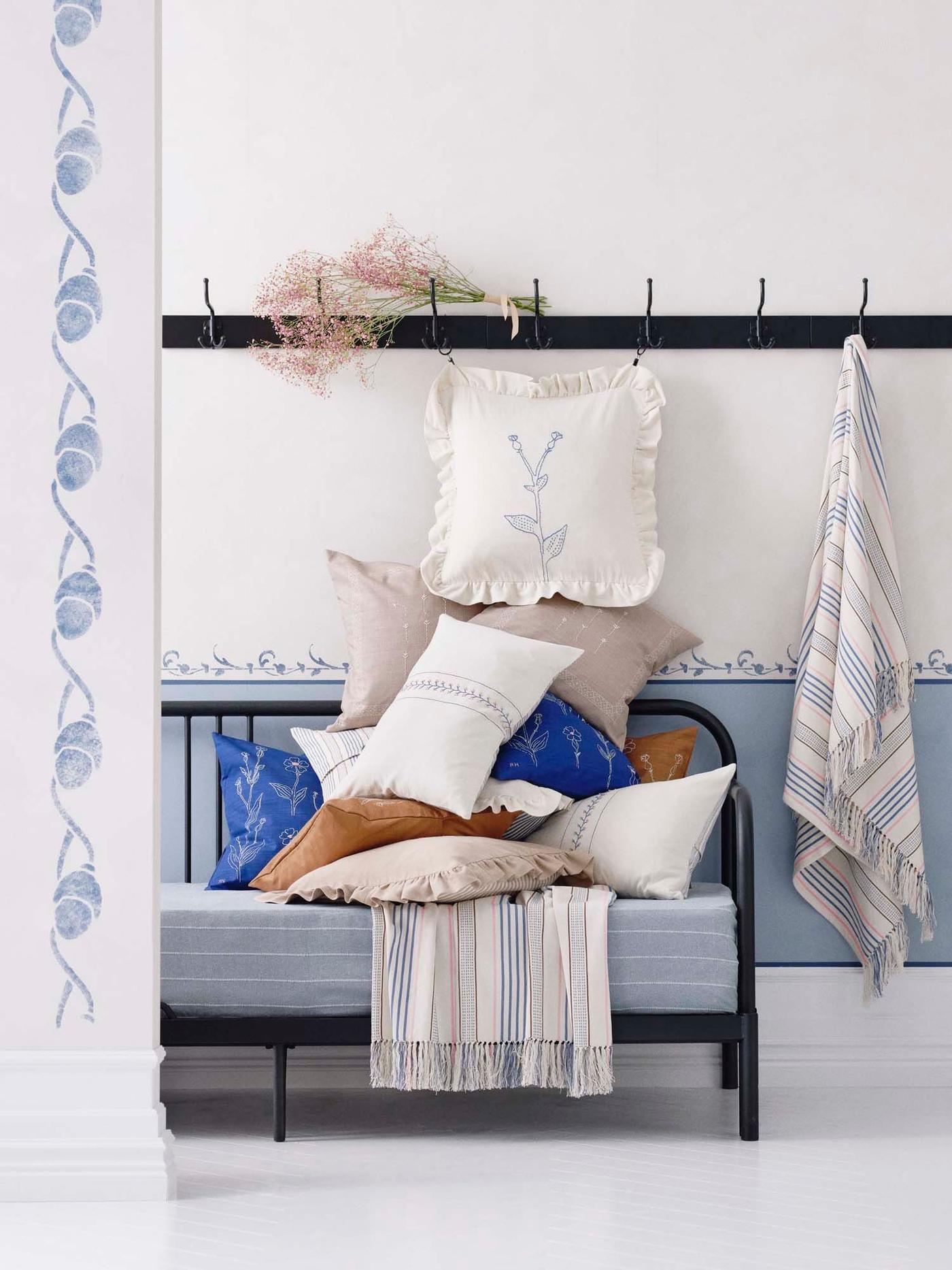 A black metal daybed is stacked with embroidered cushions and throws from the ÅKERNEJLIKA collection.