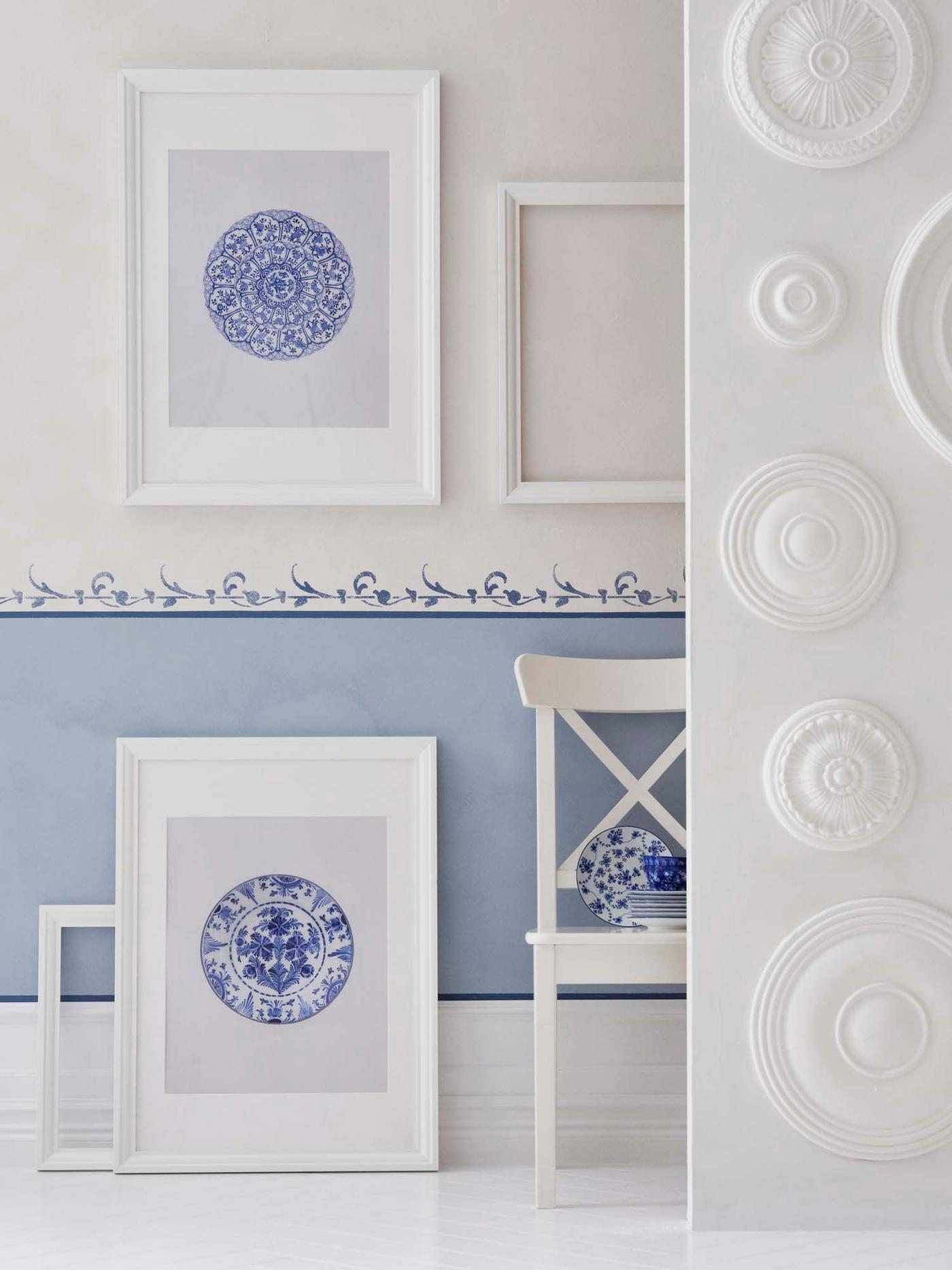 White frames and white framed images of plates hang from and lean against a blue and white wall.