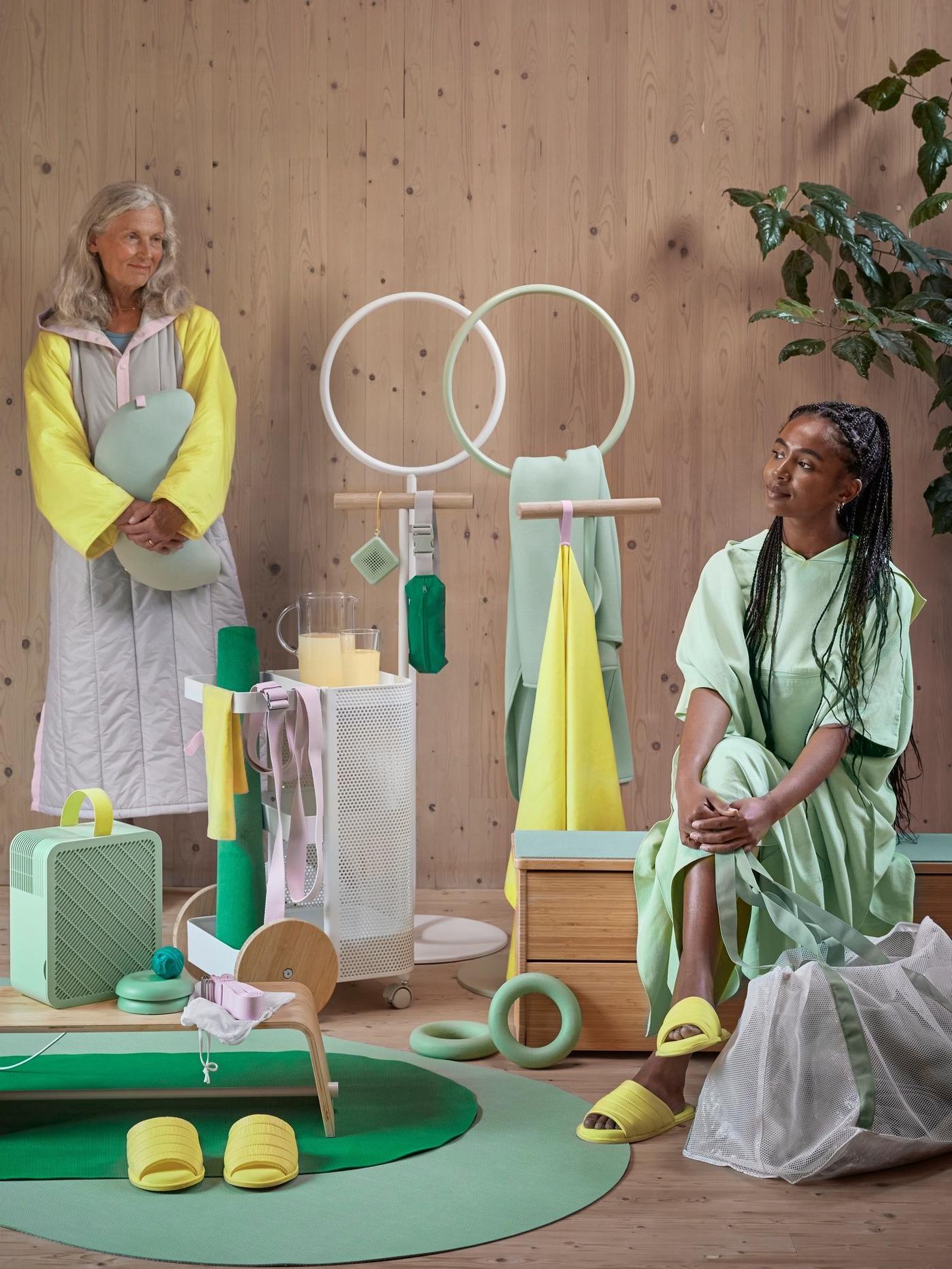 Two women in a room with items for home workouts in shades of green and yellow. Mats, towels, weights, robes, slippers, bag, clothing stands and a step up bench are included.