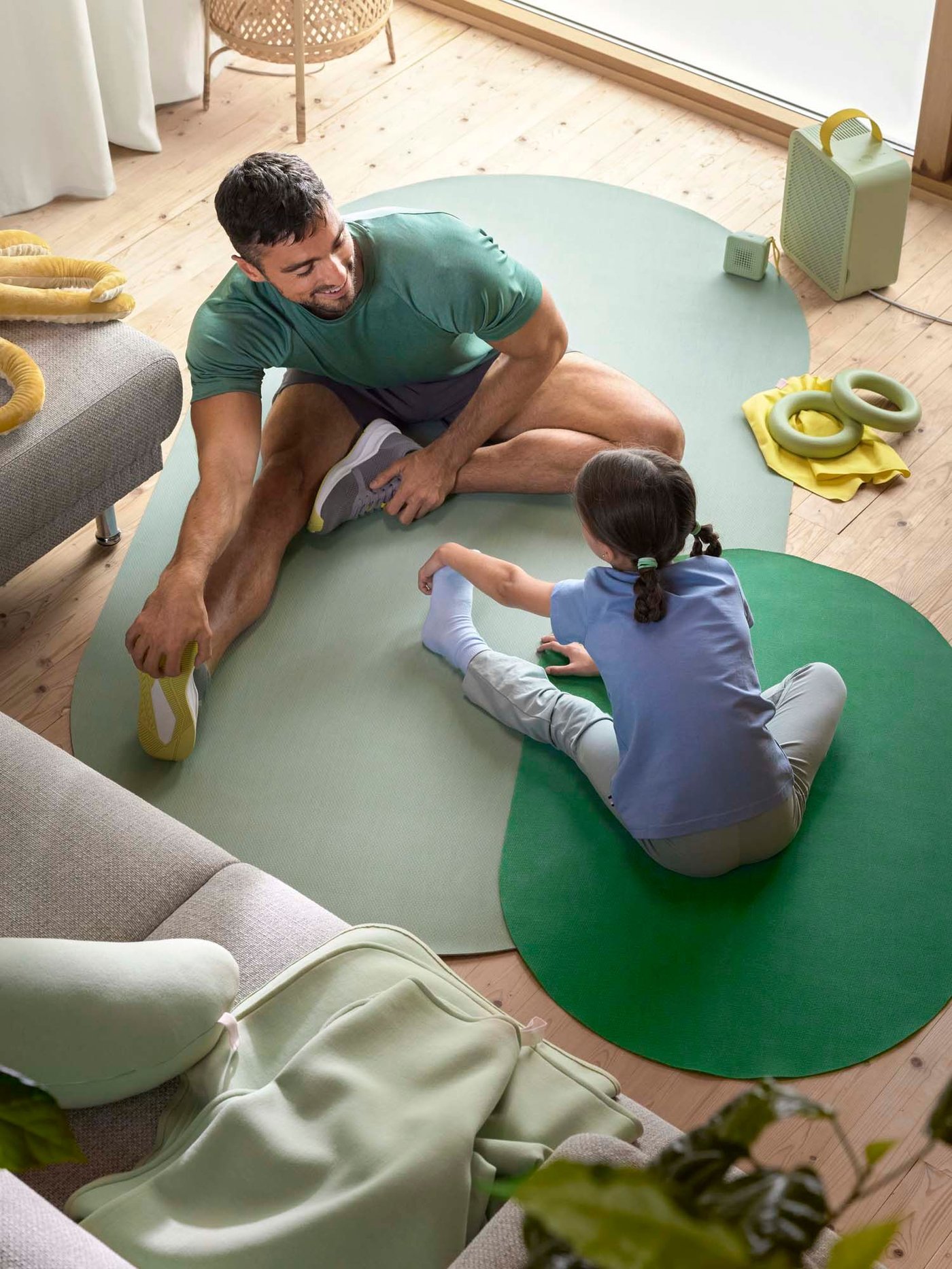 A man and child each sit on a green training mat in the living room. They are doing stretching exercises.