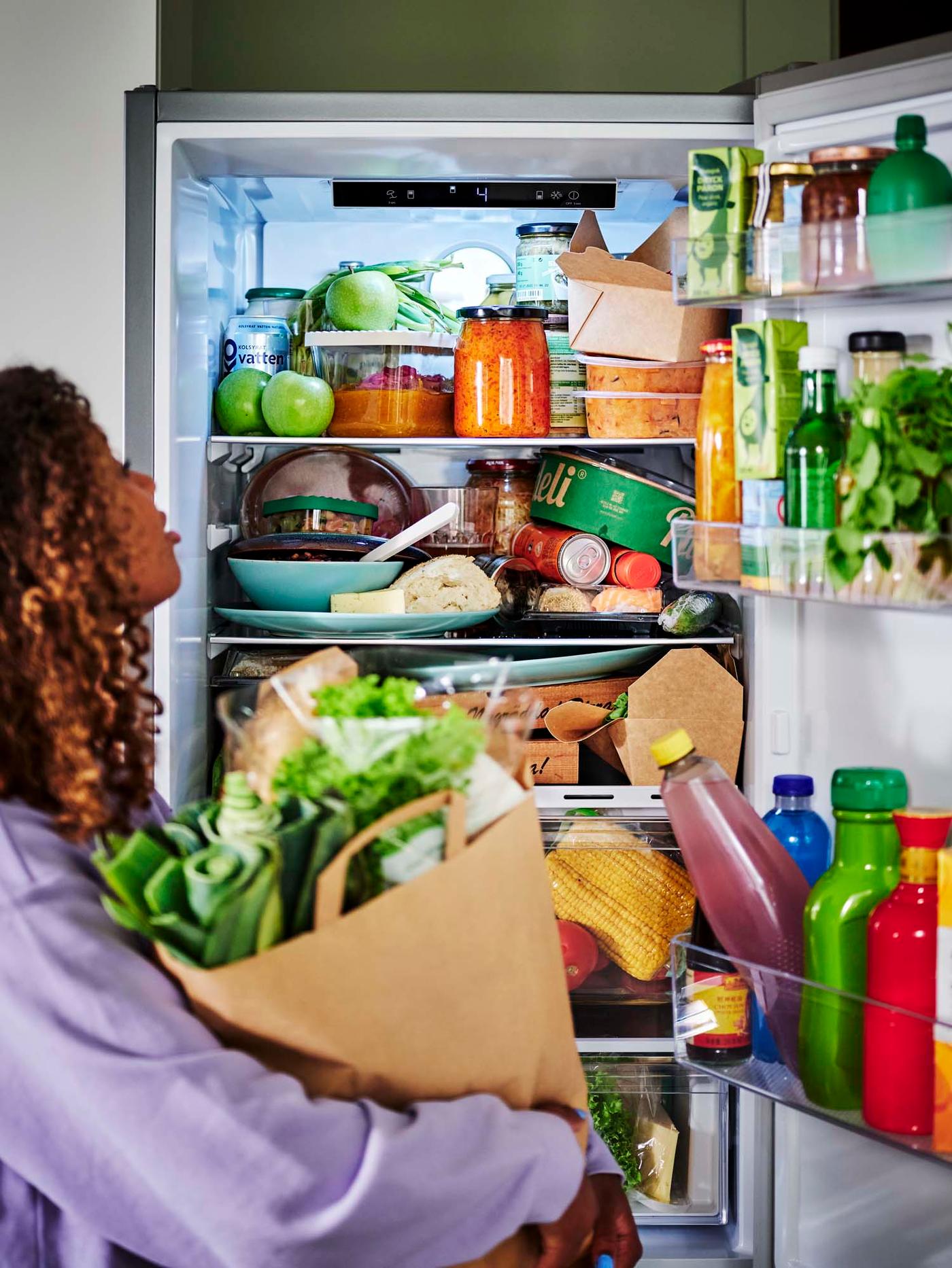 A person holding a bag of groceries stand in front of a open, well-stocked refrigerator.
