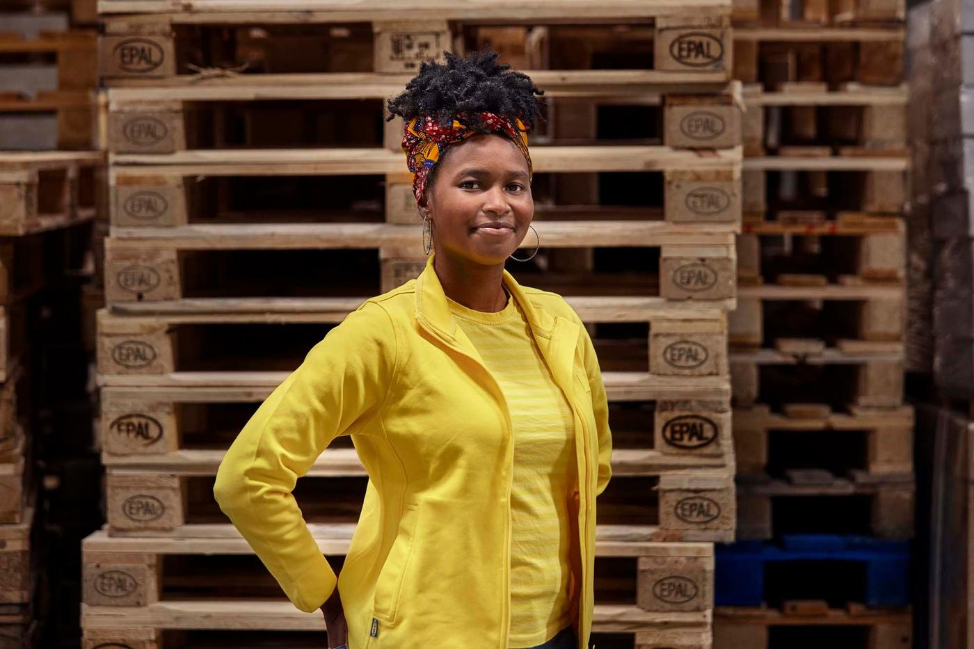 An IKEA co-worker stands in front of a stack of palettes in a warehouse, smiling at the camera.