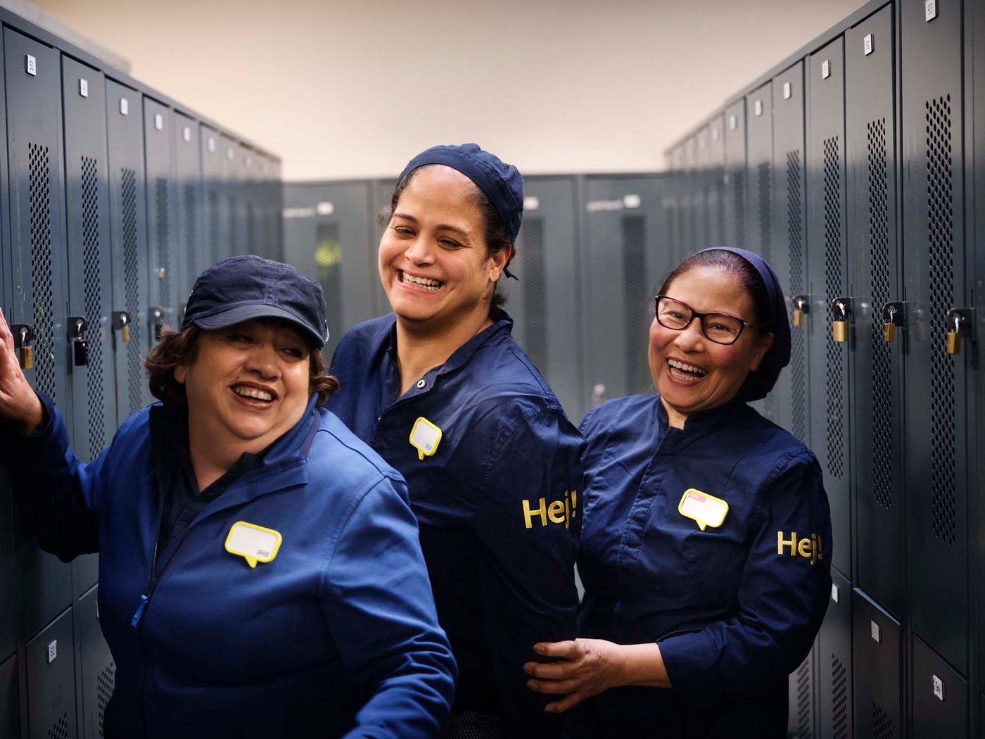 Three IKEA co-workers in restaurant uniforms stand together in a locker room. They are laughing and smiling.