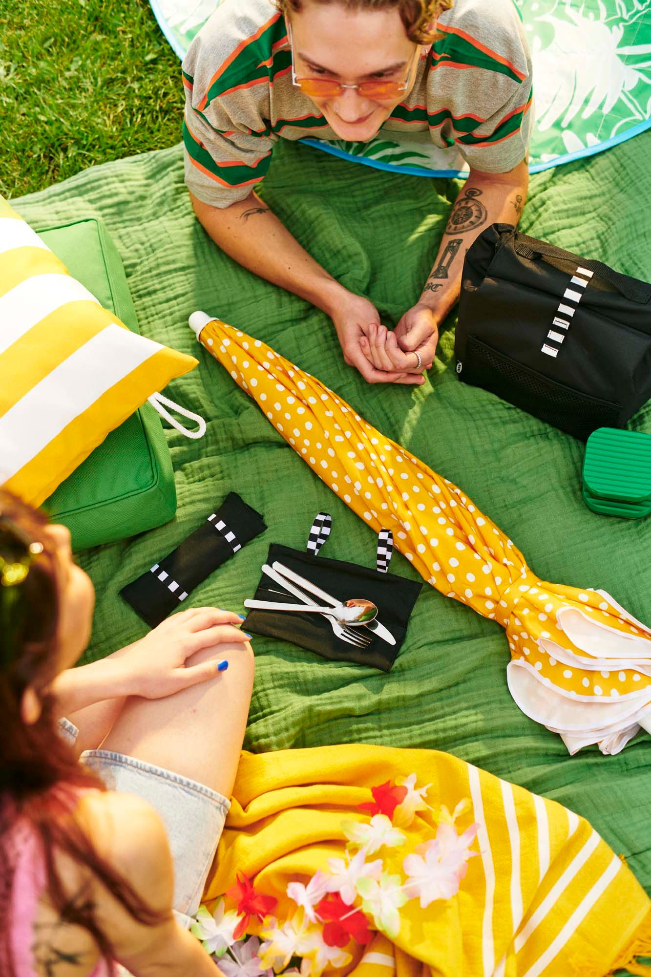 Two people picnicking on a BRÖGGAN throw with a closed yellow umbrella lying between them.
