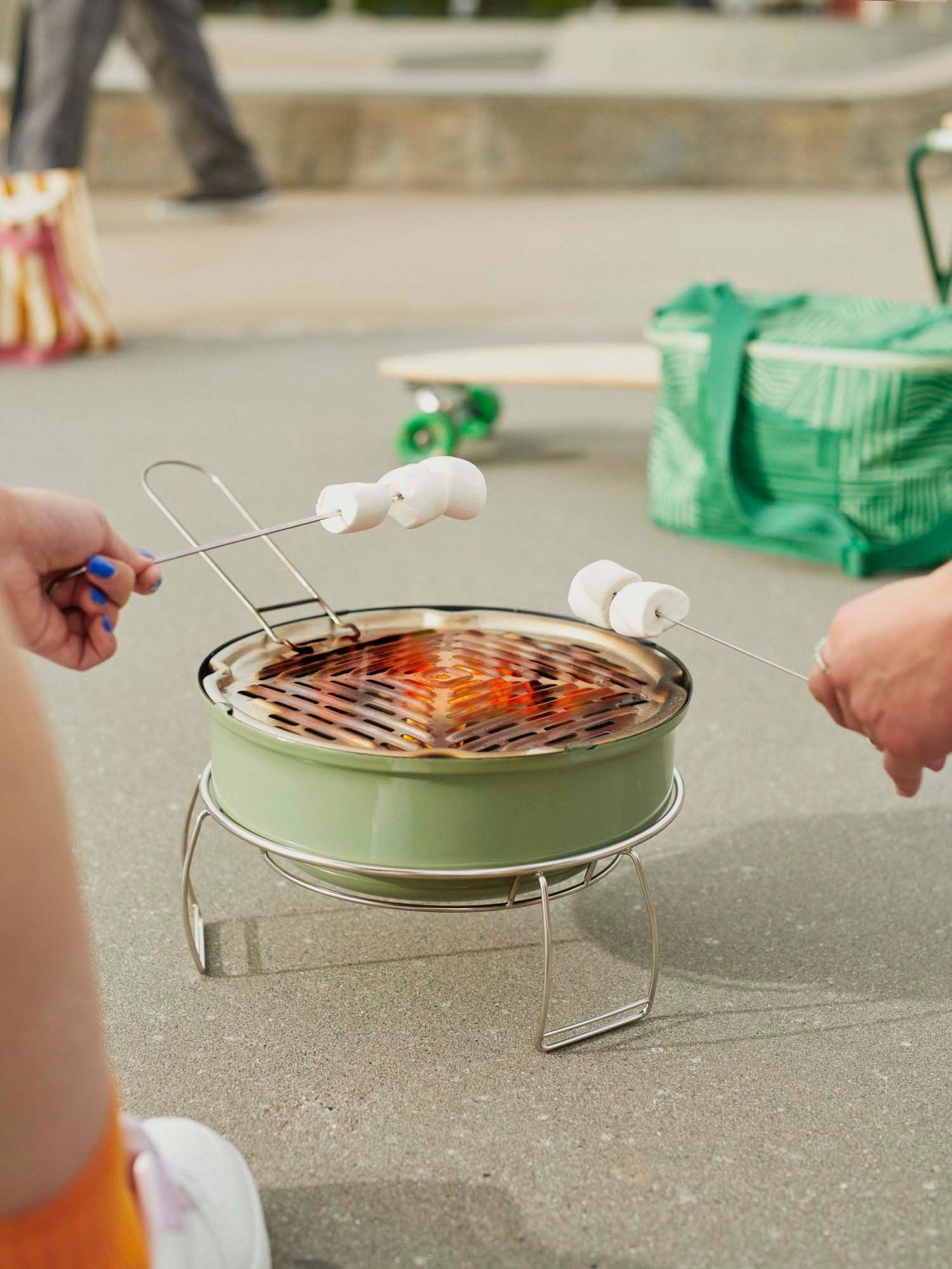 Two hands holding marshmallows over a lit STRANDÖN barbecue in a concrete environment.