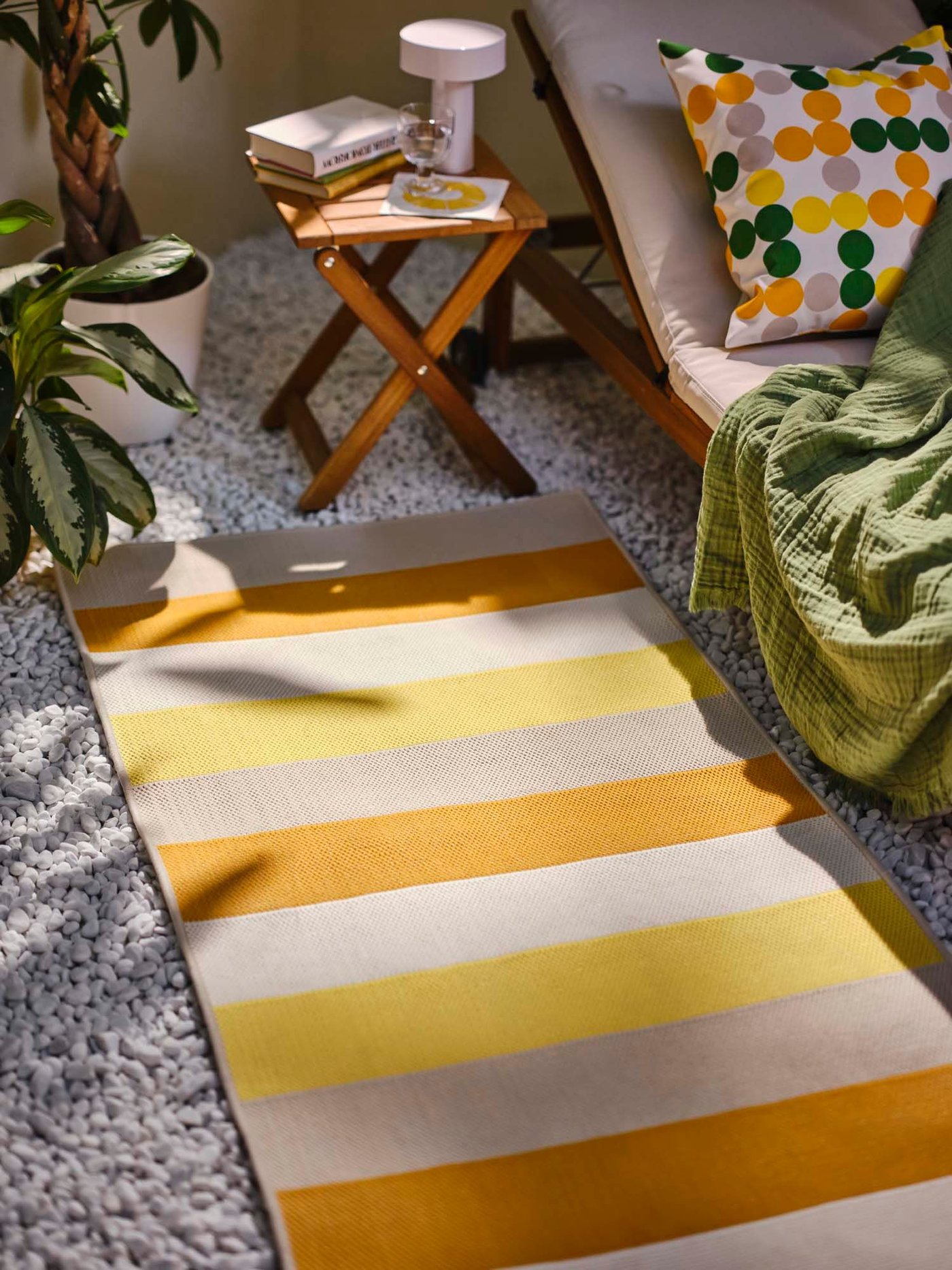 A pebbled, outdoor area with a BRÖGGAN in/outdoor rug flatwoven next to a NÄMMARÖ sun lounger and stool.