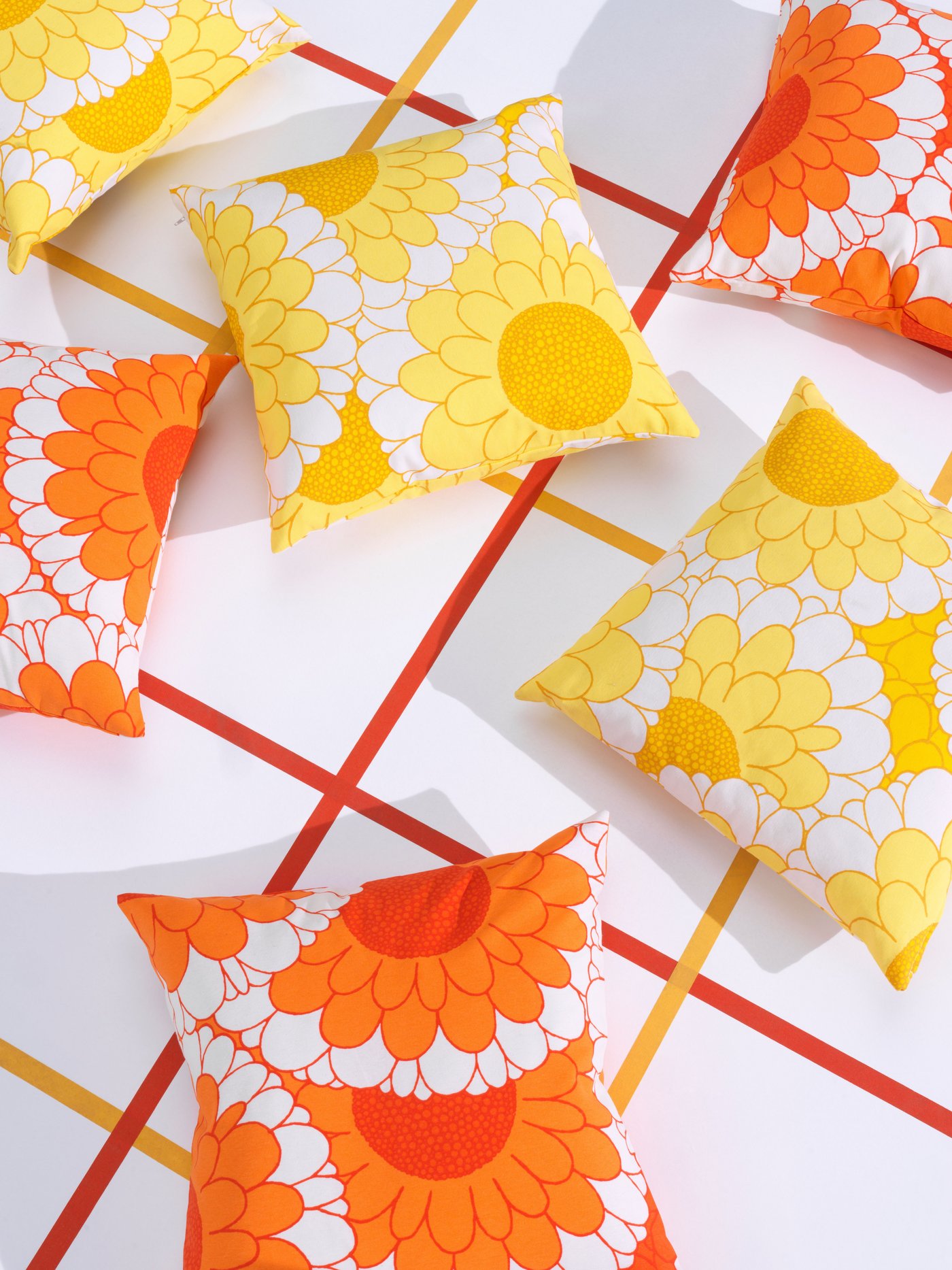 Photo with white background where orange and yellow lines are creating a block pattern. On top of it are laying 6 cushions that contain orange and yellow flower patterns.