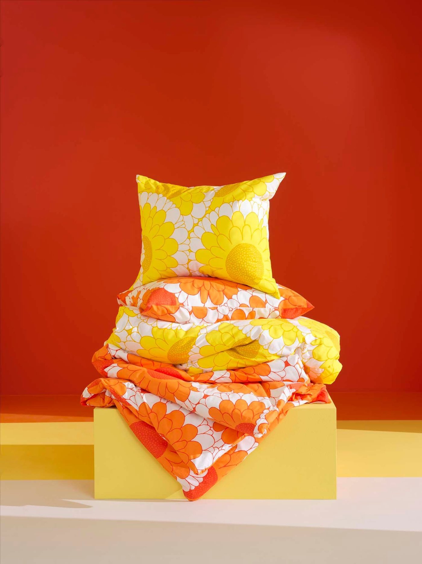 Yellow and orange KRANSMALVA bedlinen is stacked on a yellow podium, in front of an orange/red backdrop.