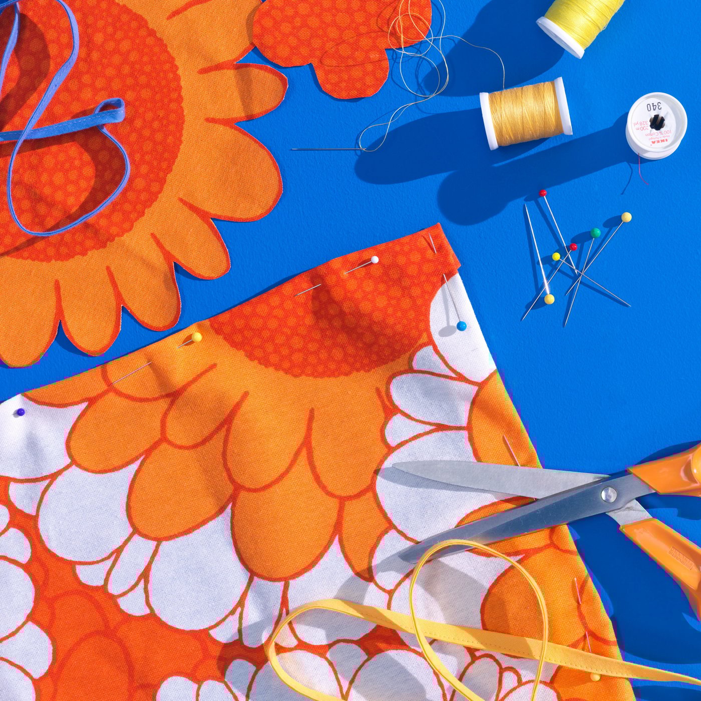 Photo of bright blue background with a colorful orange and white flower fabric. Next to it some yellow and orange yarn, scissors and needles.