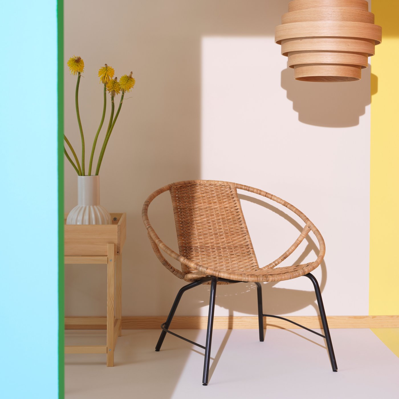 White background and floor, natural material chair and next to it a natural material table with a white vase with yellow flowers. A natural lamp hanging from ceiling.