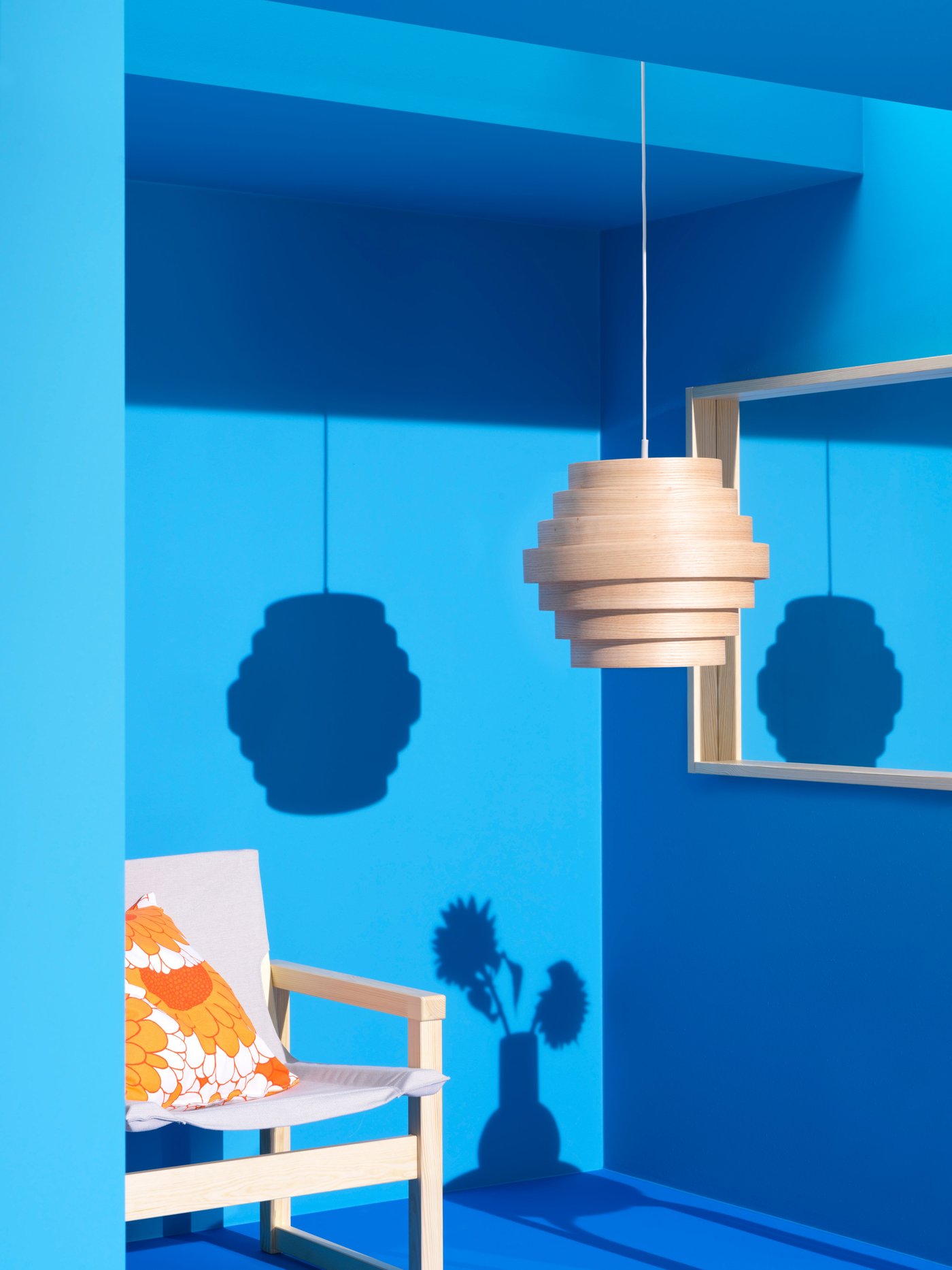 Blue background, natural/white chair with orange flower pattern cushion. From ceiling a natural lamp hanging.