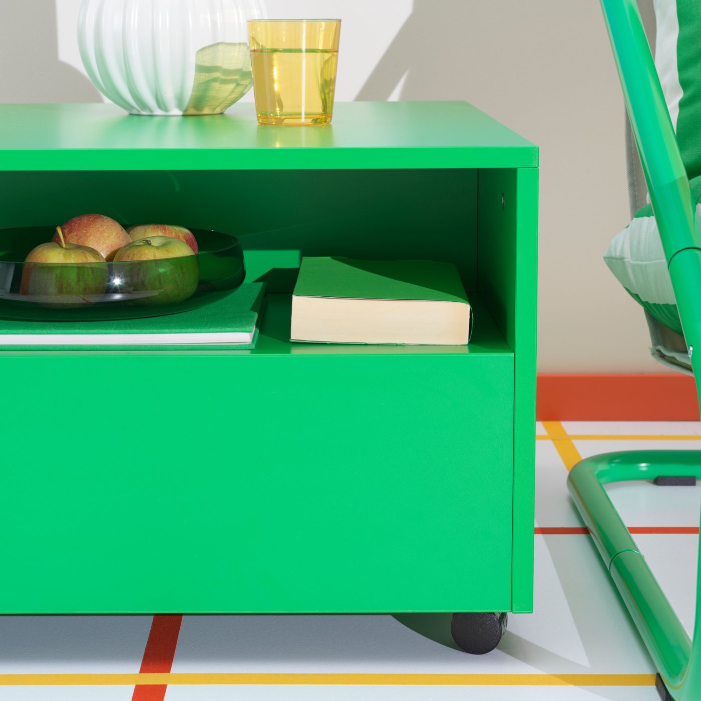 Green small dressoir with accessories on it like a yellow glass, white vase, a book and glass fruit basket with apples. 