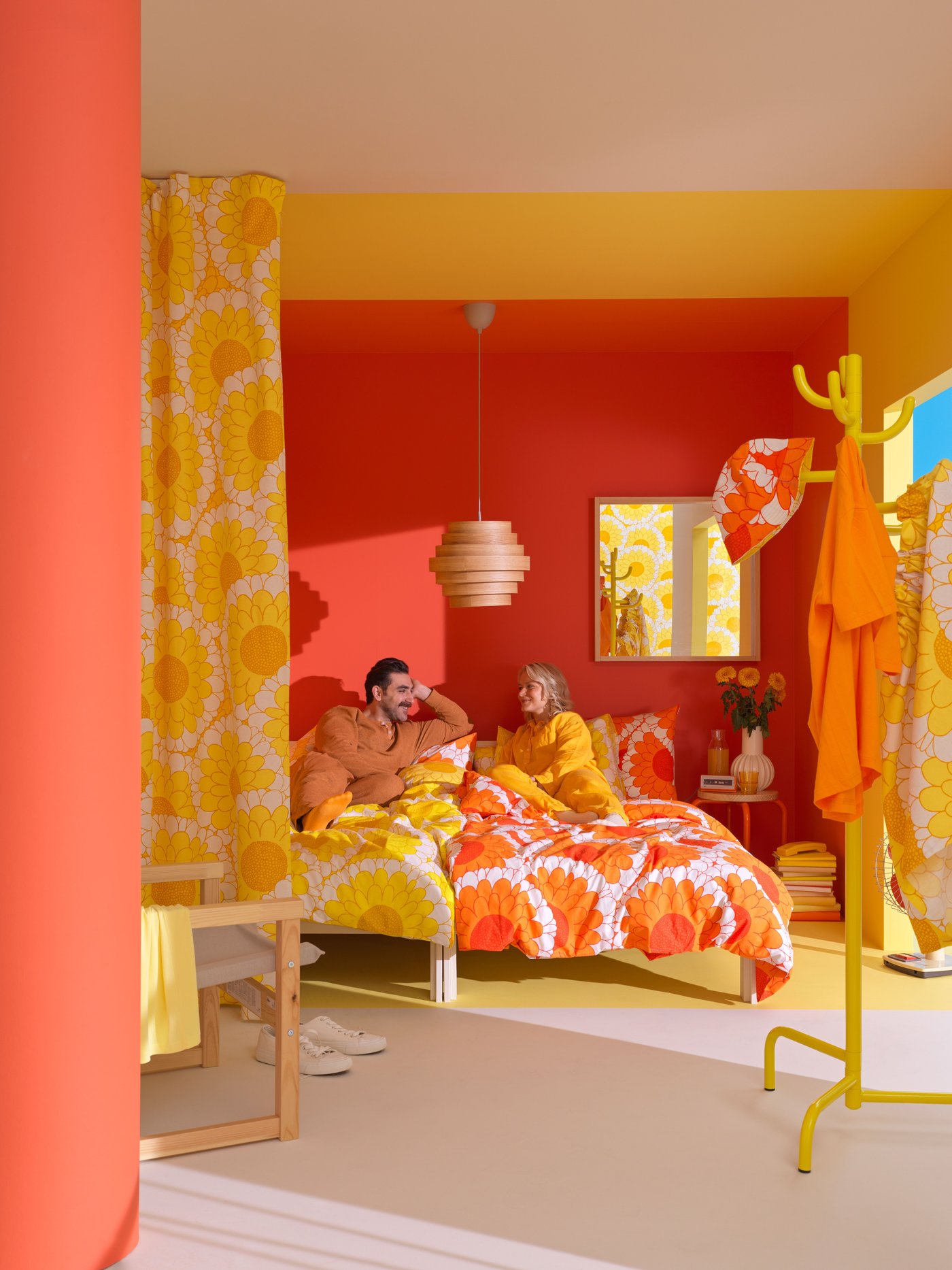 A man and woman in orange and yellow pyjama's laying on bed with floral pattern bedsheets, in a yellow and orange room 