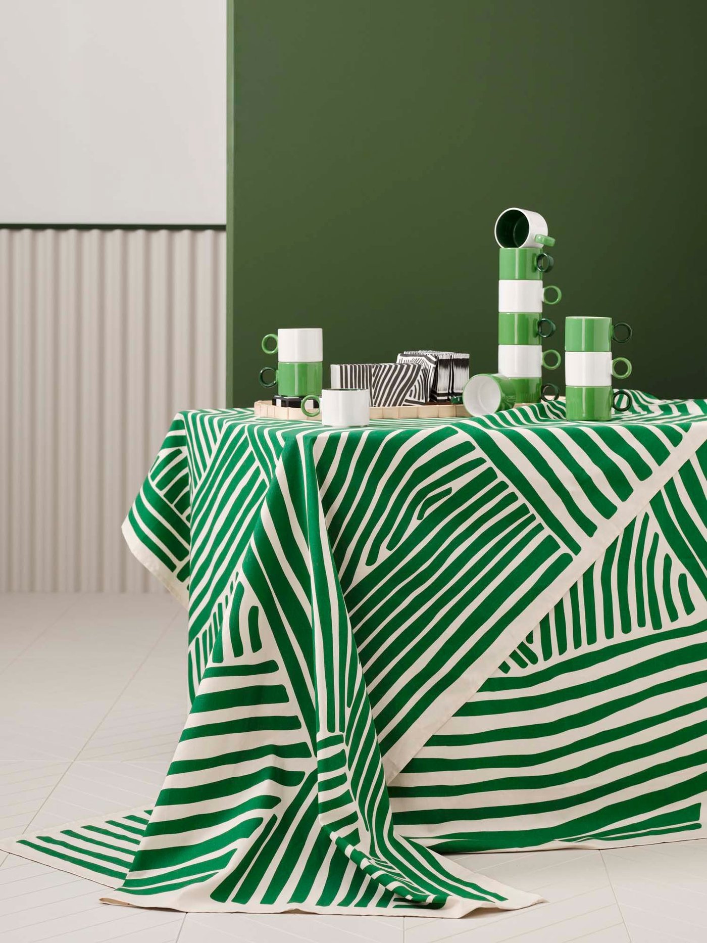 A table with a cream and white striped tablecloth and green and white cups, stands in front of a green wall.