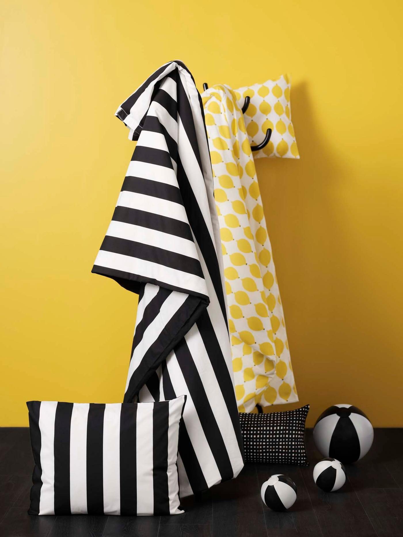 A yellow wall with yellow and white patterned and black and white striped linen hanging from hooks. There's a striped pillow and three balls on the floor.