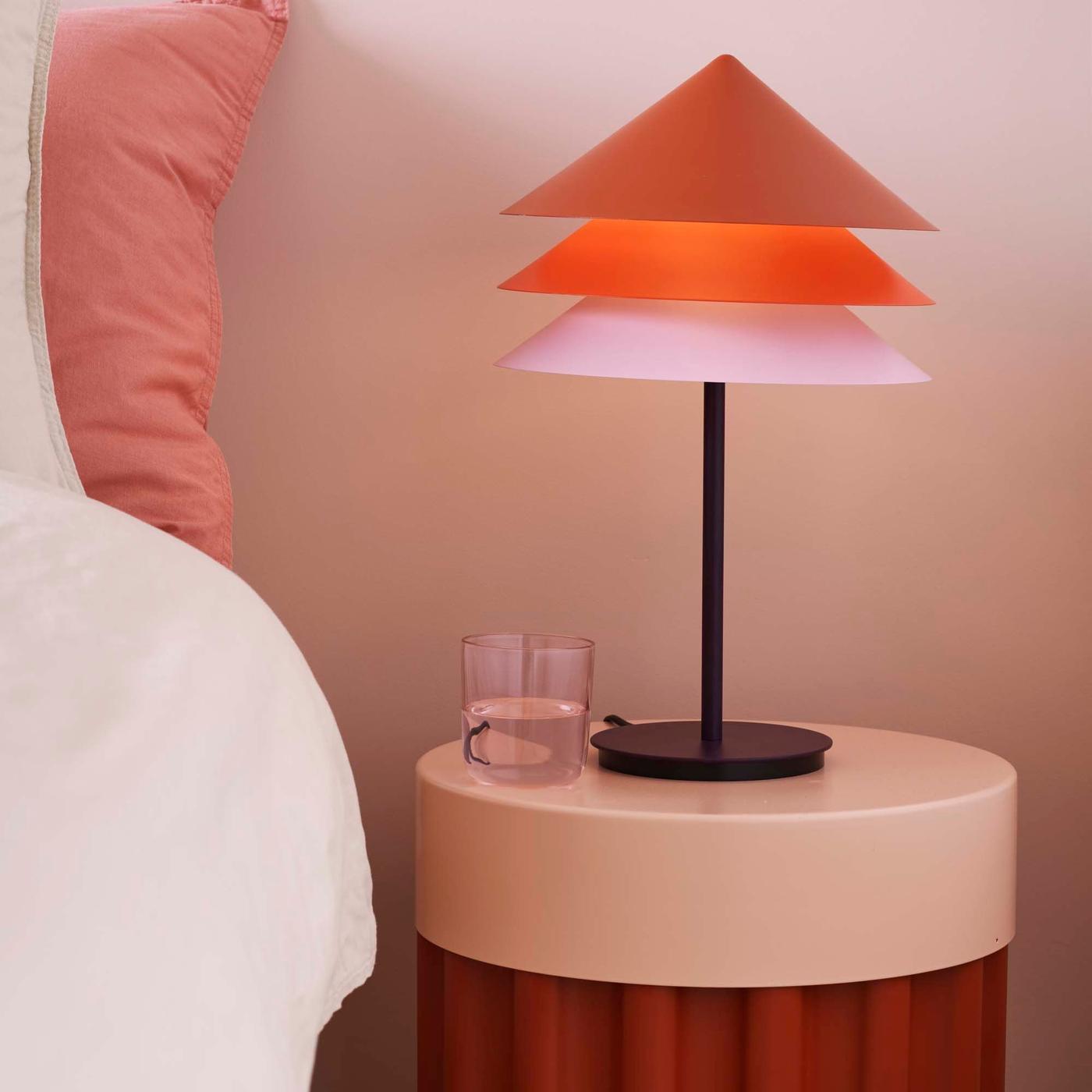 A lit lamp with a TESAMMANS lampshade atop a TESAMMANS side table, next to a bed.
