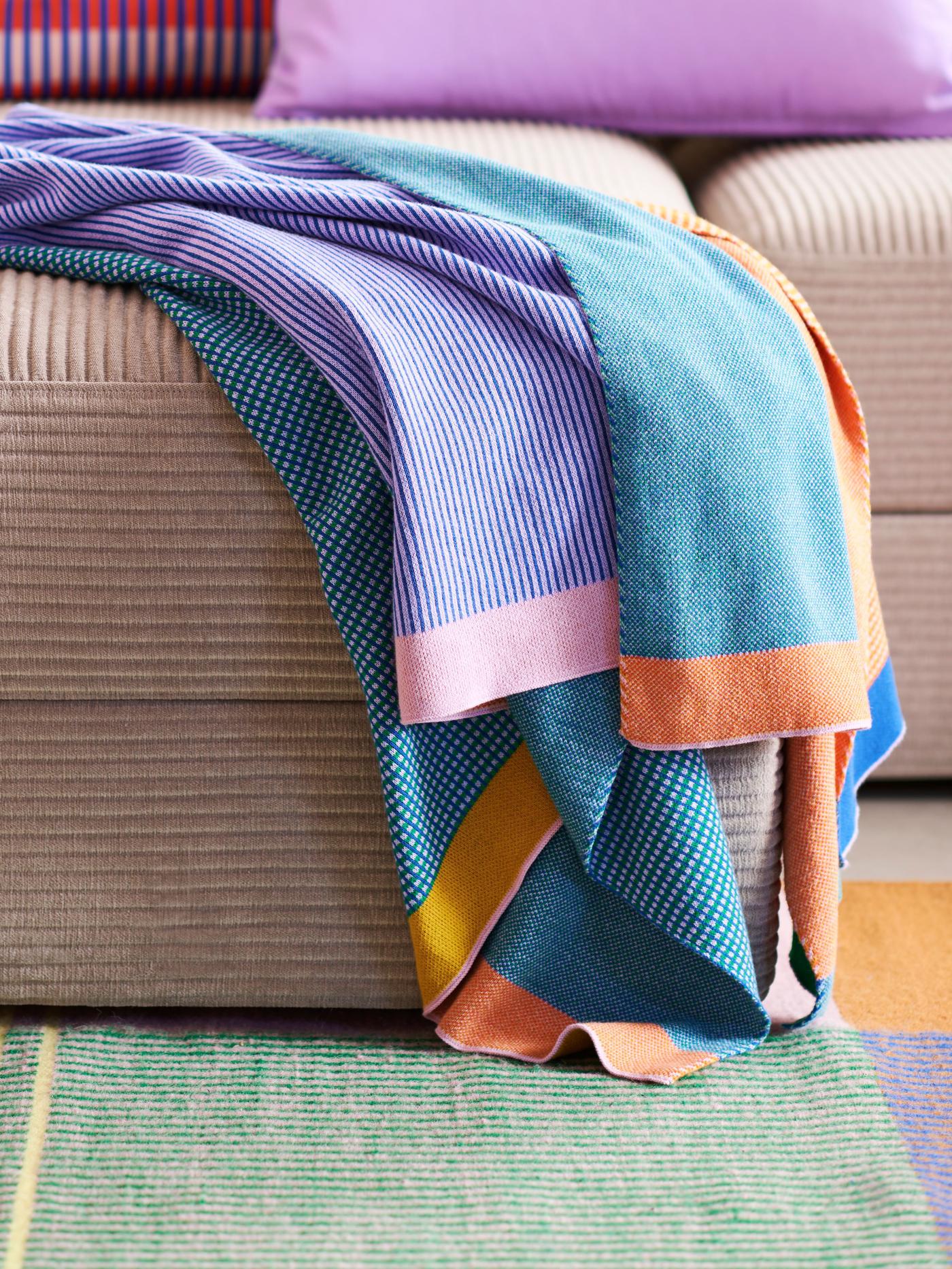 A knitted throw of blue, purple, pink, yellow and orange is folded and lying on a beige sofa