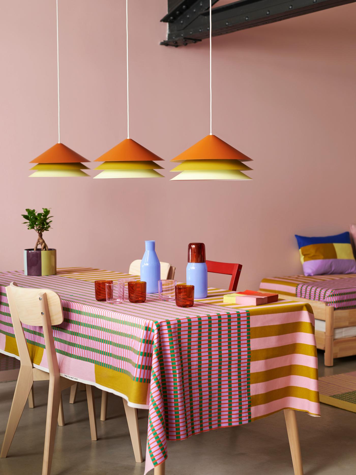 Three lampshades are hanging from the ceiling over a dinner table made with blue vases, red glasses with a colourfully striped cloth over it.