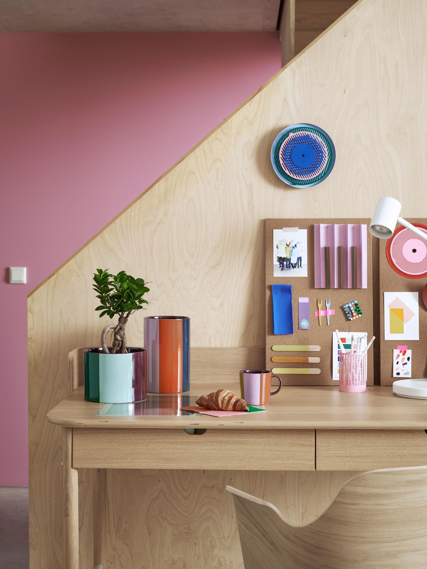 A clock of different colours, flower pots and a coffee cup are standing on a wooden workspace desk in front of a pink wall.