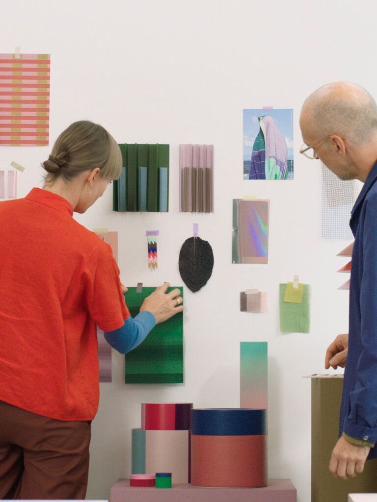 Daniera ter Haar and Christoph Brach of Raw Color stand with their backs against camera, looking towards and touching sketches of different sizes, shapes and forms that are attached to the wall. Next to it are several prototypes.
