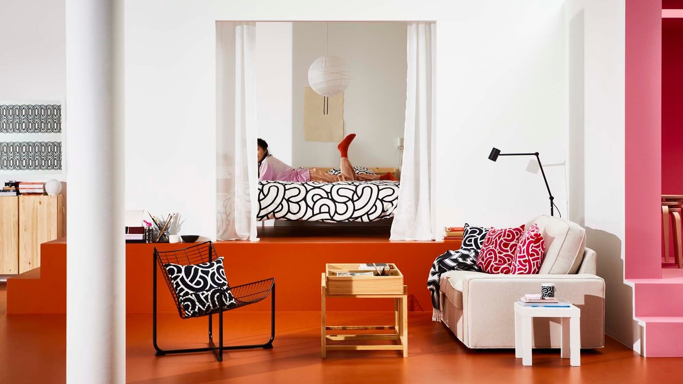 A room with a sofa, chair and table. In the background, a person is lying on the bed in the bedroom.