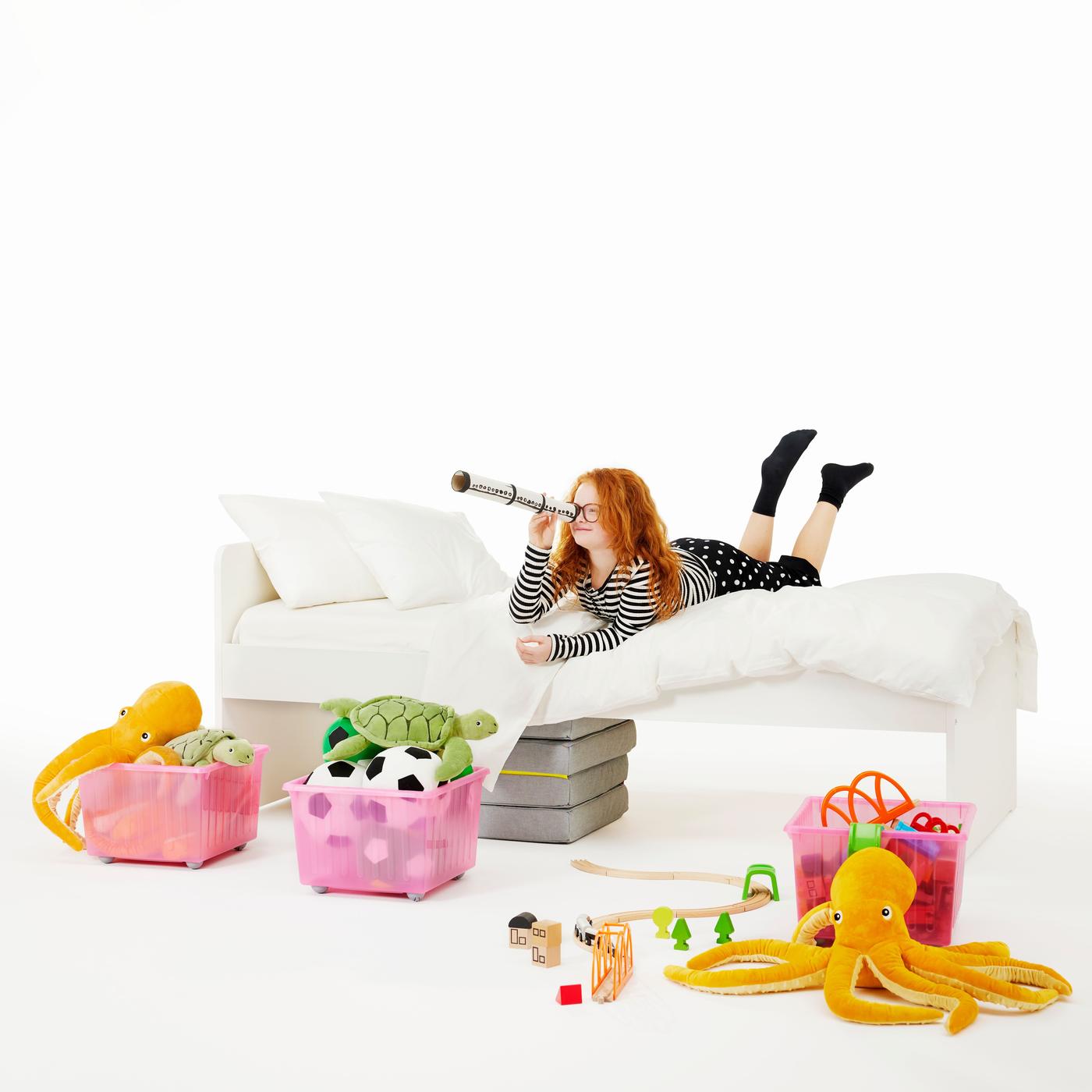 A girl laying on a white bed, holding a small telescope and looking through it. Around her on the ground several colorful boxes and buckets with IKEA soft toys. 