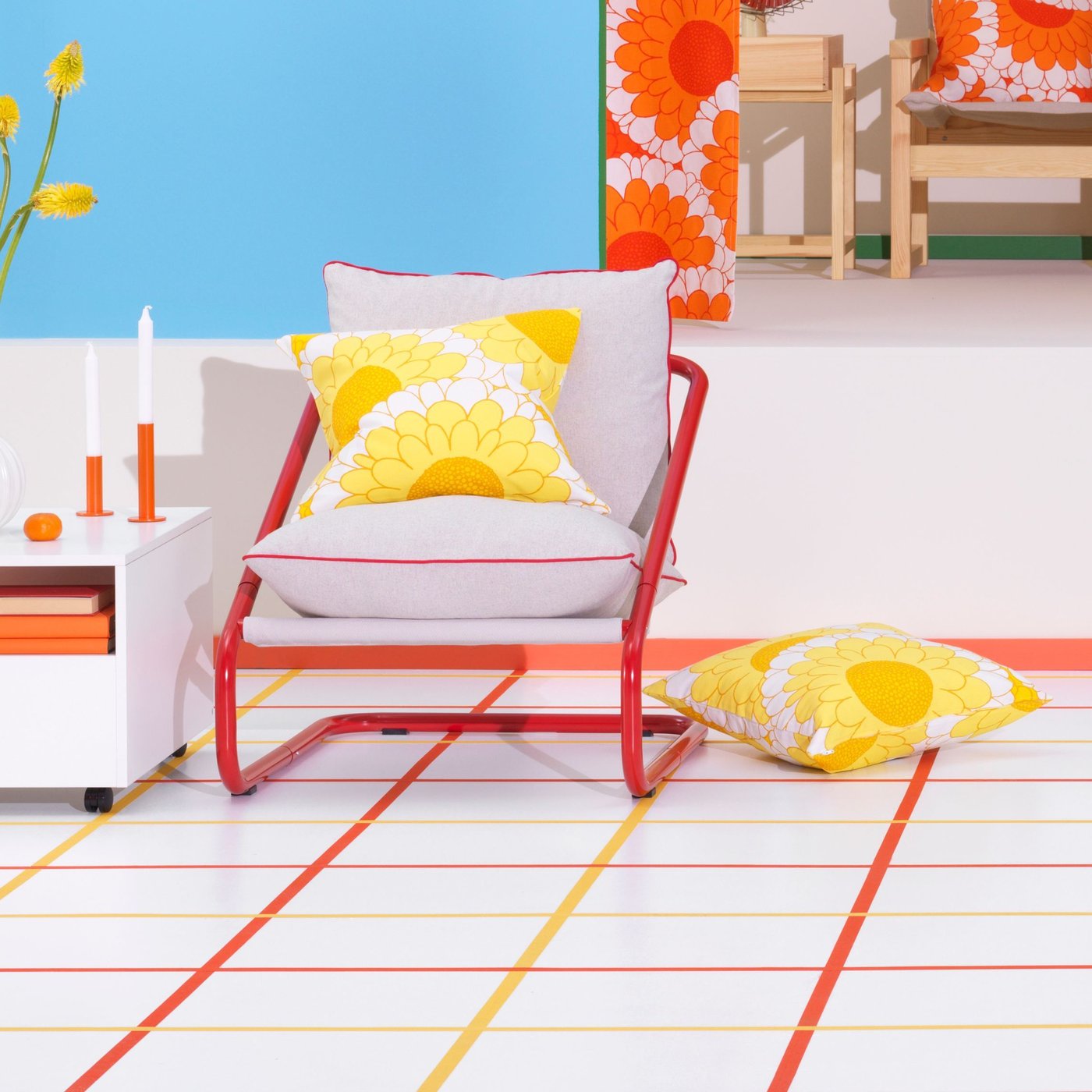 Photo with white floor with orange and yellow lines creating a block pattern. Red and beige chair with a yellow flower pattern cushions. Next to the chair a white table with accessories. Background a blue wall and orange flower patterns.
