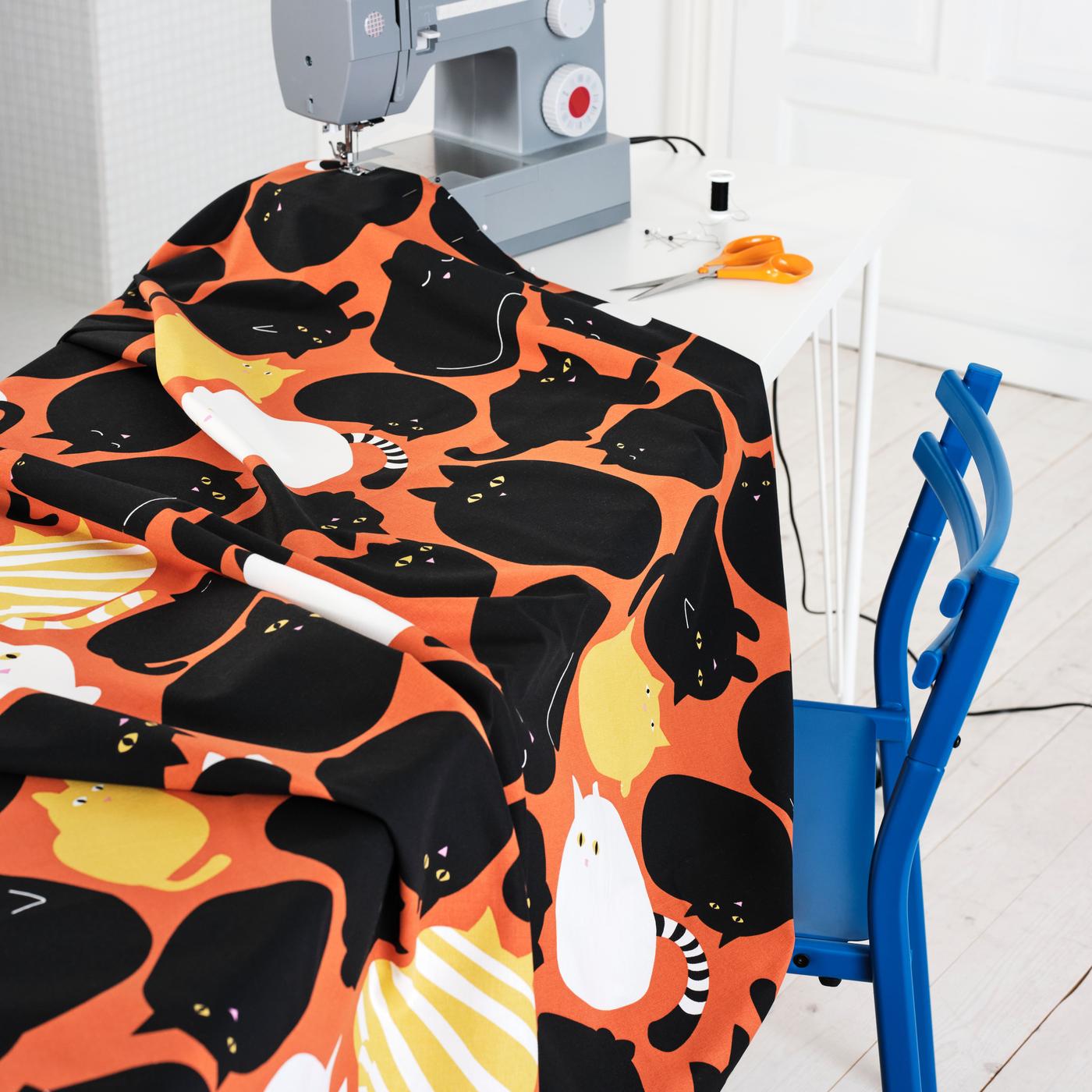 On the desk is a sewing machine, and by the side is a meter of fabric with cat patterns. Besides the table, there is a chair. 