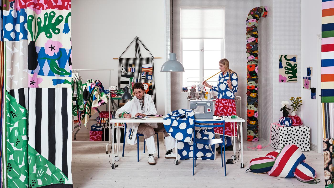 Two people are sitting and standing in a spacious room, with various decorations made and covered with patterned fabrics. 