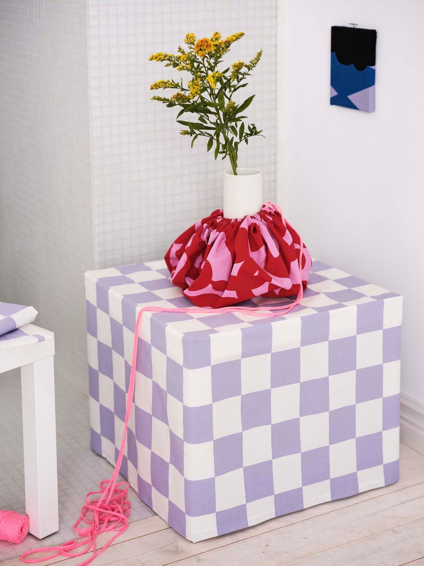 A vase covered in a handmade pink and red cover stands on a small table, which is covered in lilac chequerboard fabric.